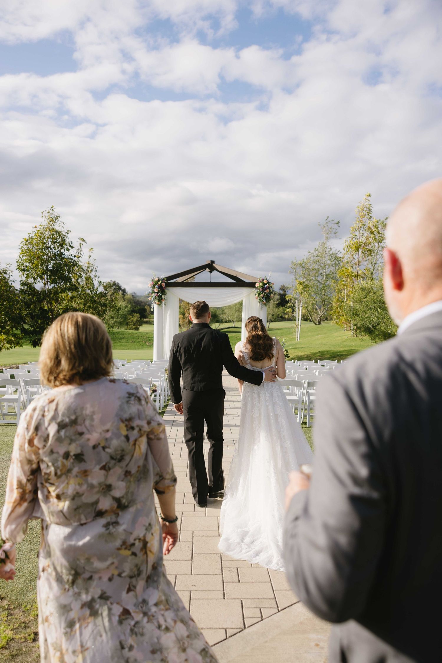 Wedding guests walk up a stone path toward a white outdoor ceremony gazebo on a cloudy summer day.
