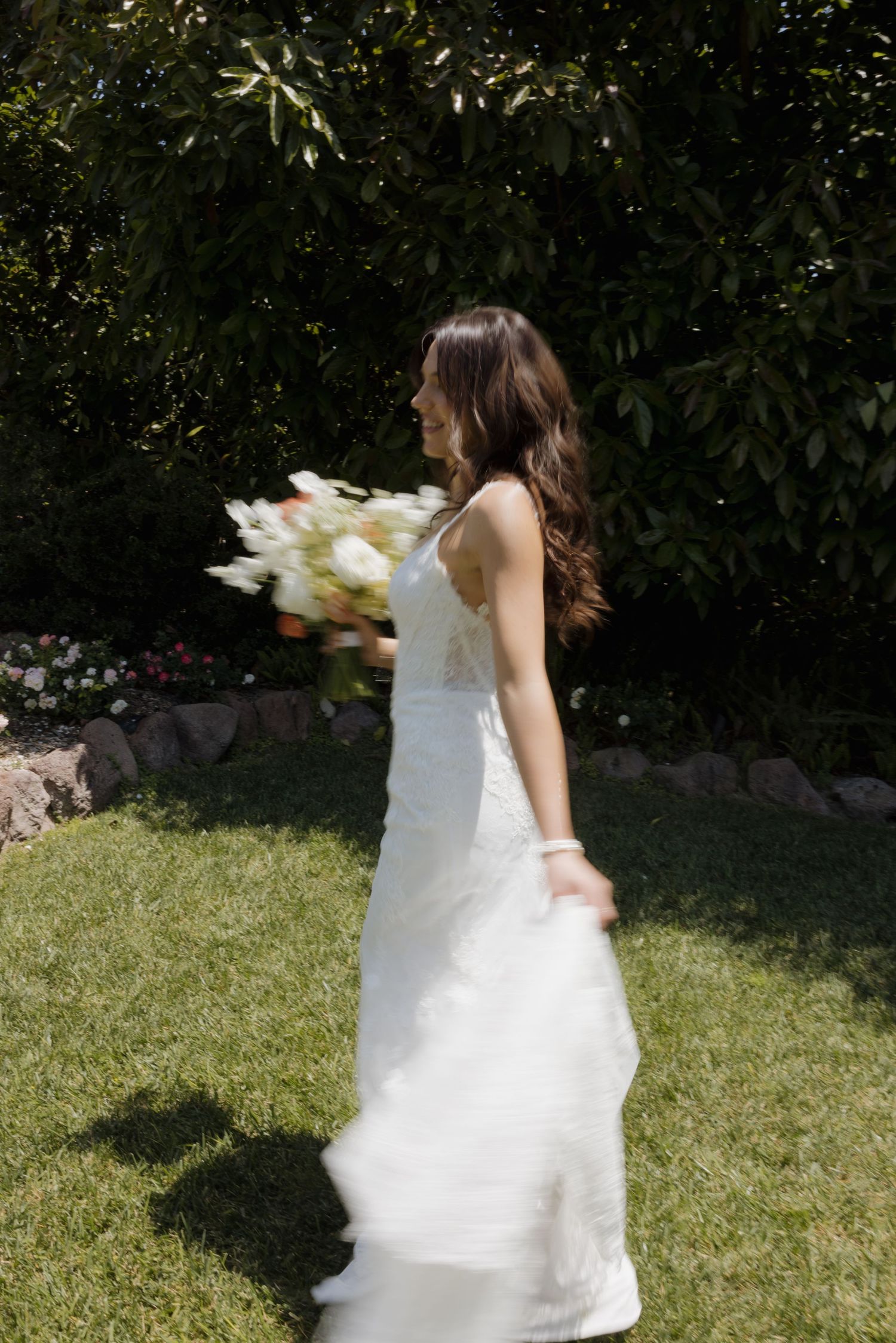 Bride in white gown holding white tulip bouquet walking on green grass in sunlit garden.