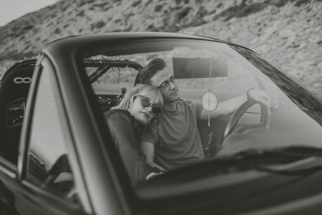 Black and white photo of two people sitting in car against mountain backdrop.