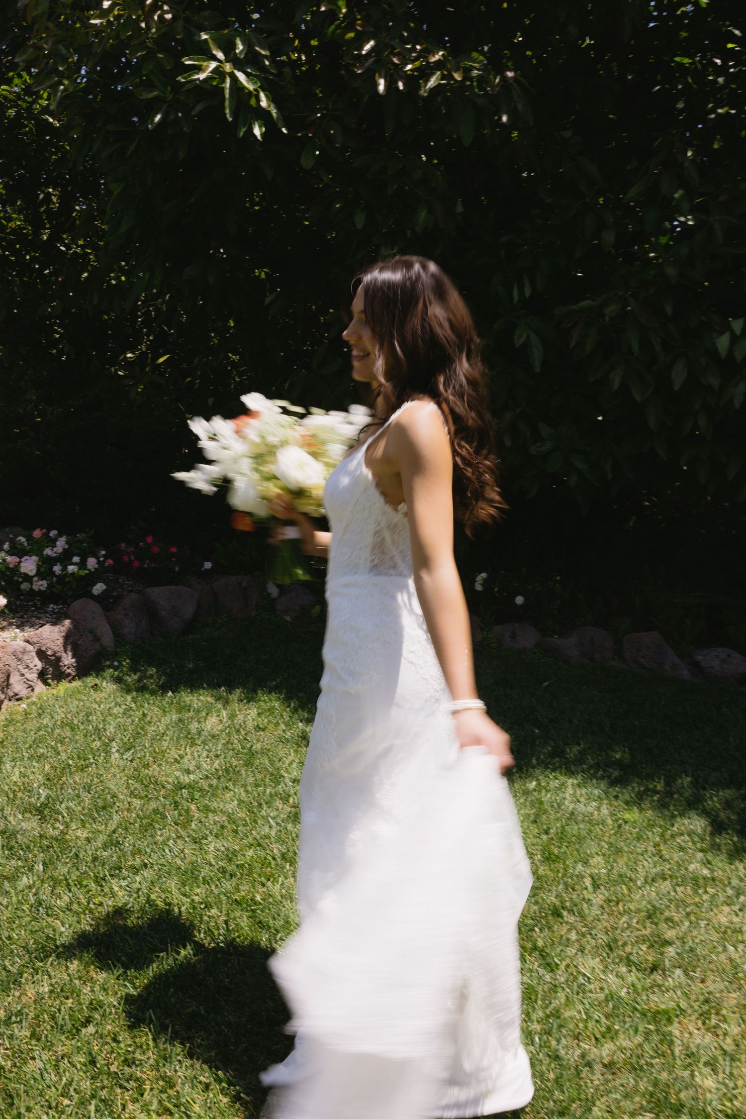 Bride in white gown holding white floral bouquet walking on green grass with lush garden background.