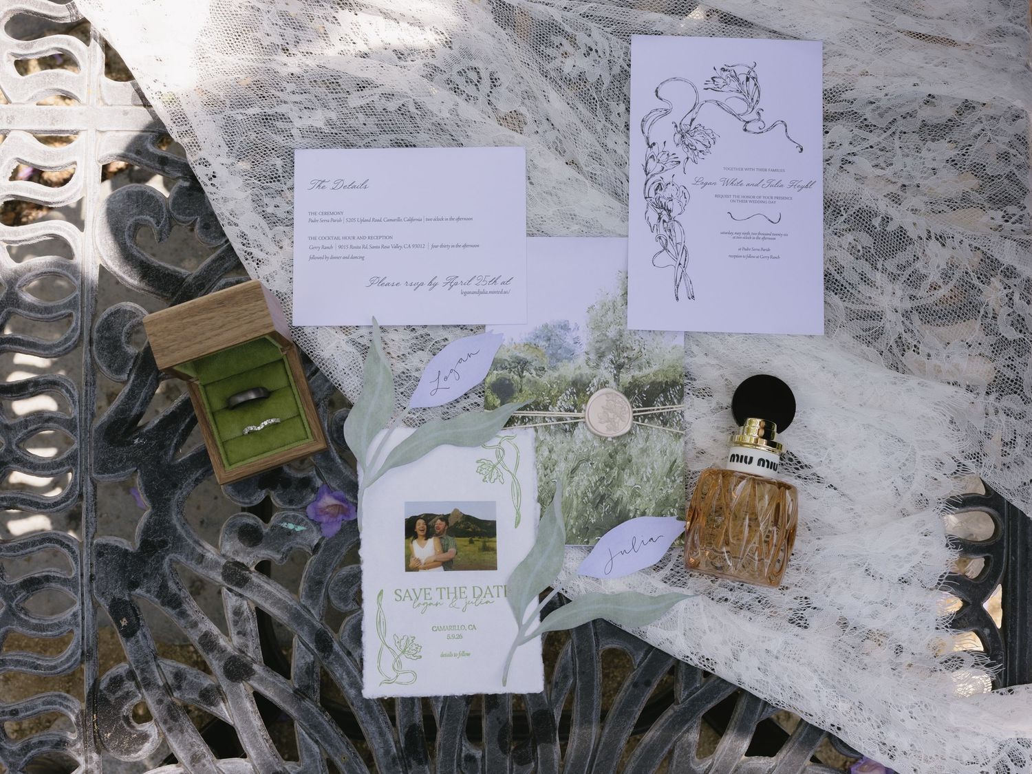 Elegant wedding stationery flat lay with invitations, save the date, rings, and perfume on lace.