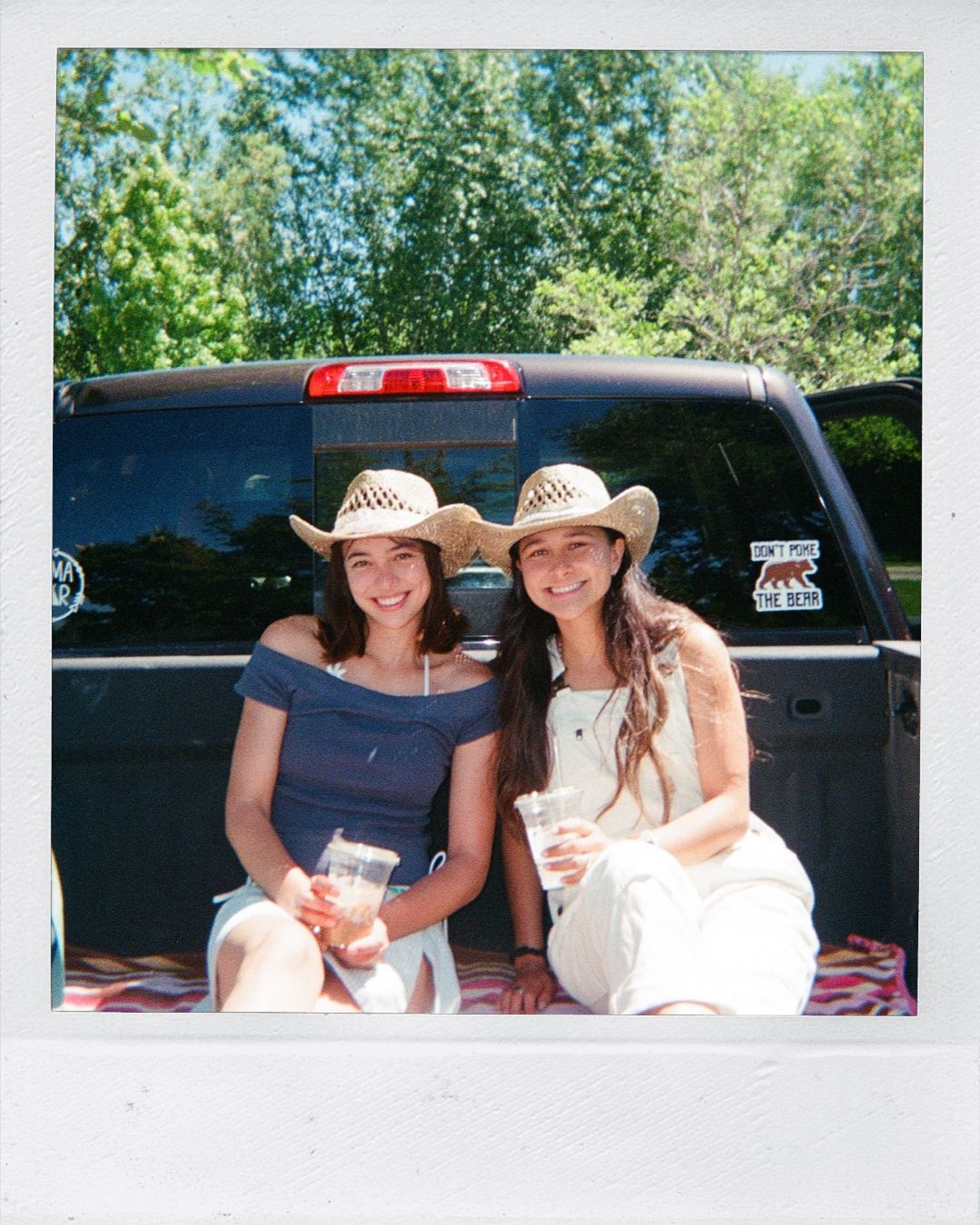 Two smiling women in cowboy hats sitting in a truck bed on a sunny day, holding drinks outdoors.