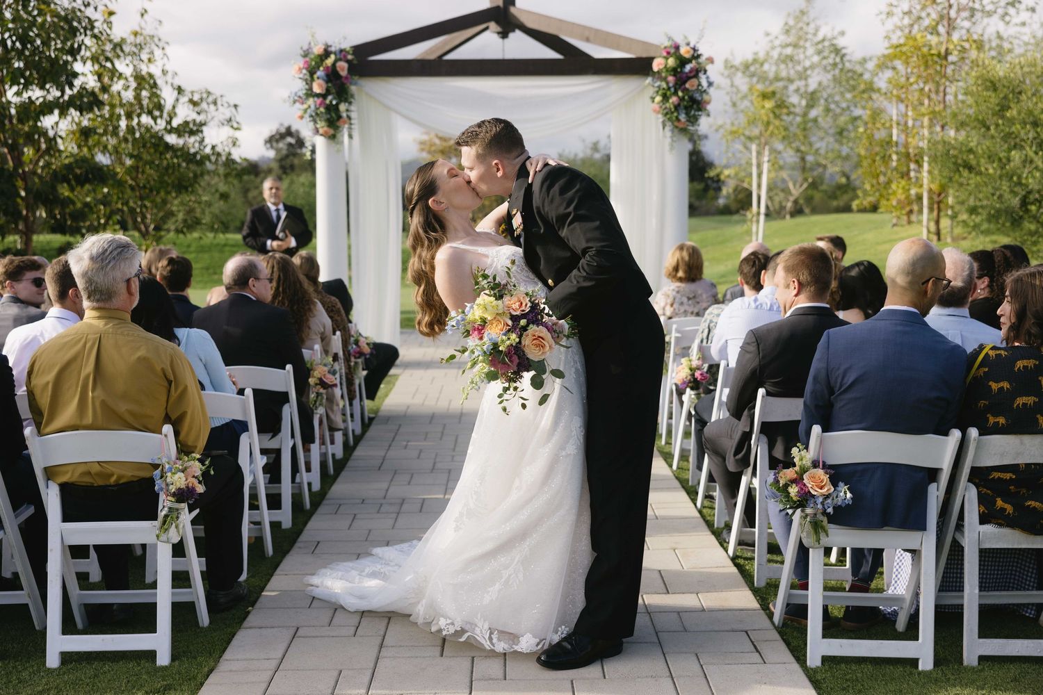 Newlyweds share a romantic kiss under a floral arch during an outdoor garden ceremony with guests seated on white chairs.