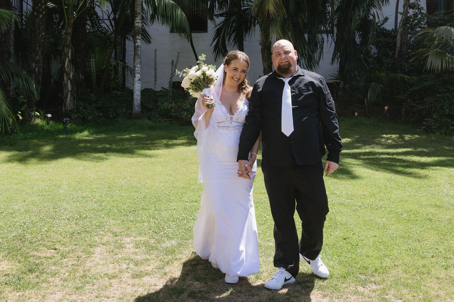 A couple in wedding attire standing together on a lawn with palm trees in the background.