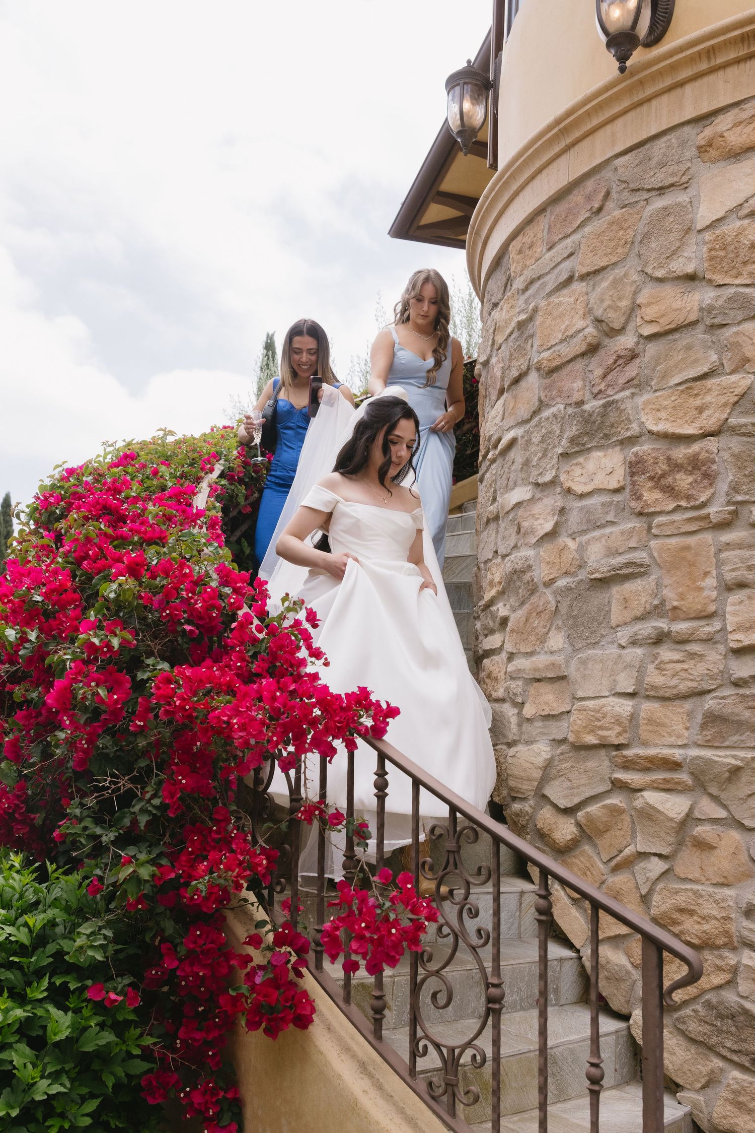 Three women descending stone staircase with vibrant red bougainvillea flowers along wrought iron railing.