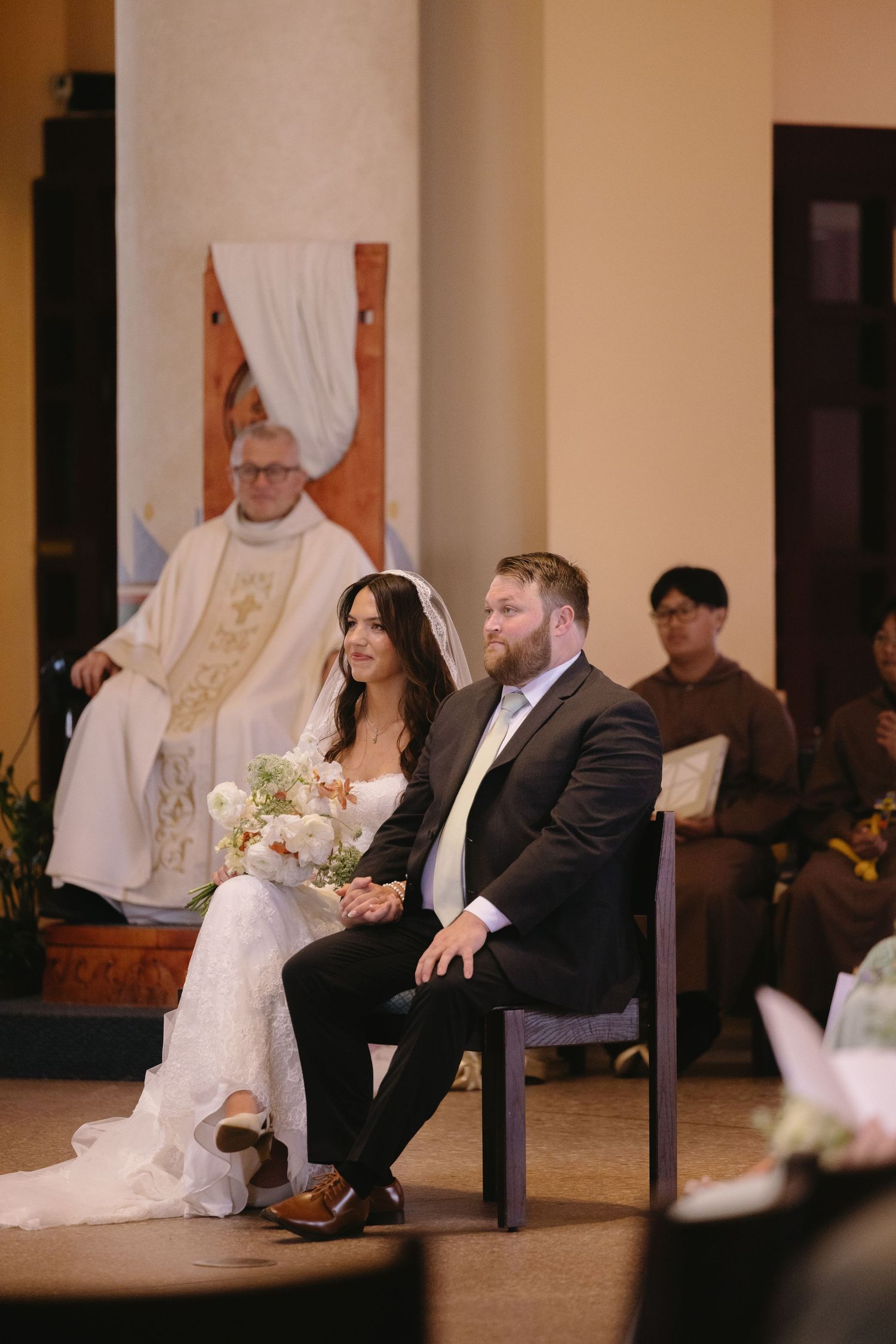 Bride and groom seated at church altar during wedding ceremony with priest officiating in white vestments.