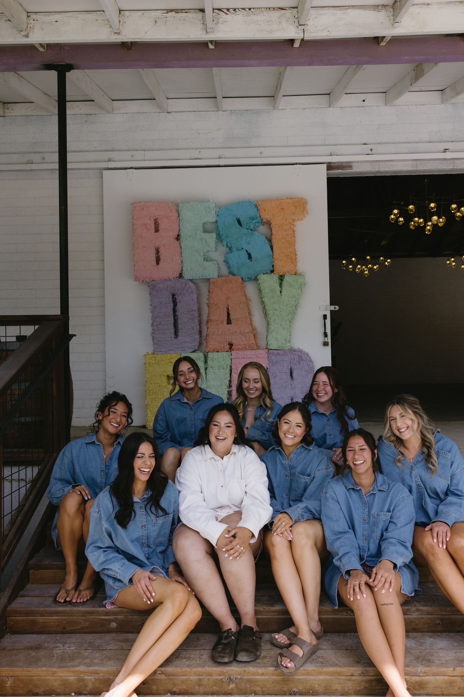 Group of bridesmaids in denim shirts sitting on steps in front of colorful Best Day Ever sign.