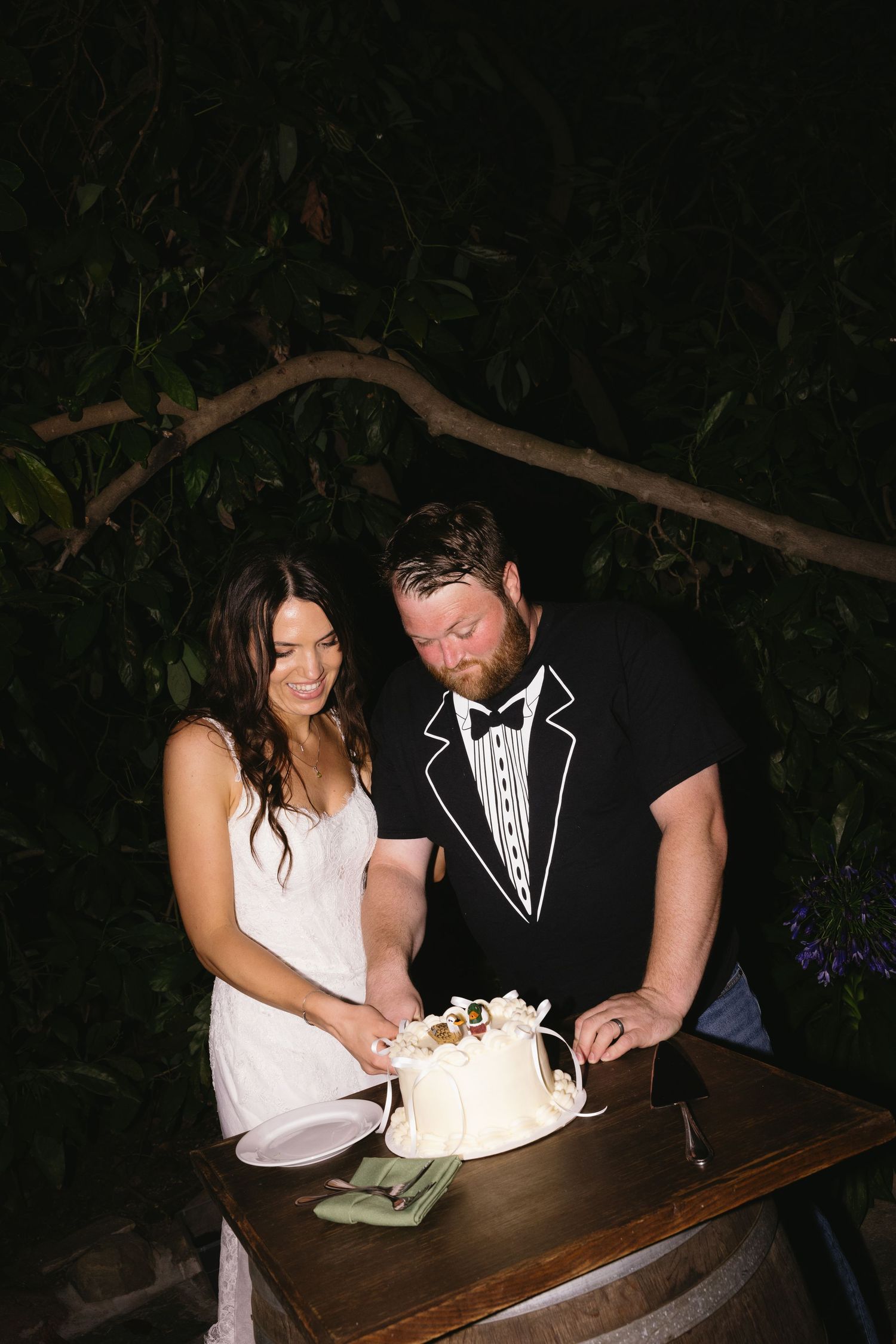 Bride and groom cutting white wedding cake outdoors at night, man wearing tuxedo t-shirt, woman in white dress.