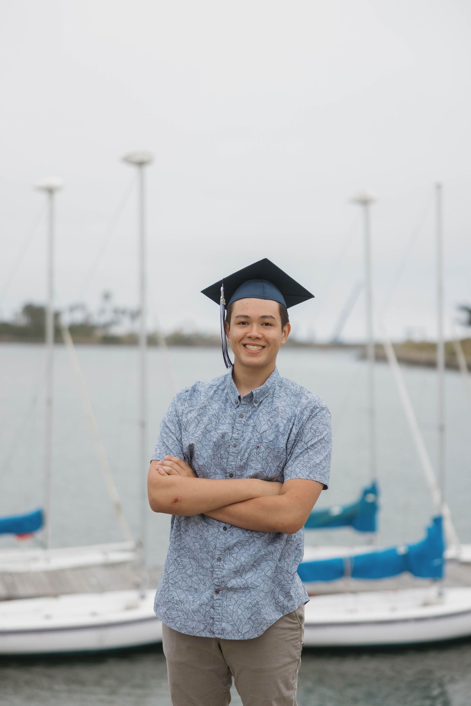 A graduate in a black mortarboard cap stands with crossed arms by a marina with sailboats in the background.