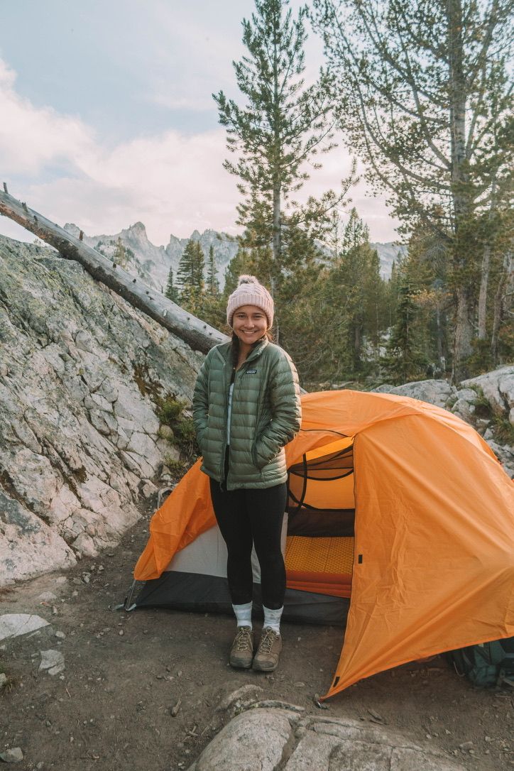 A winter camper stands by an orange tent in a snowy mountain setting.