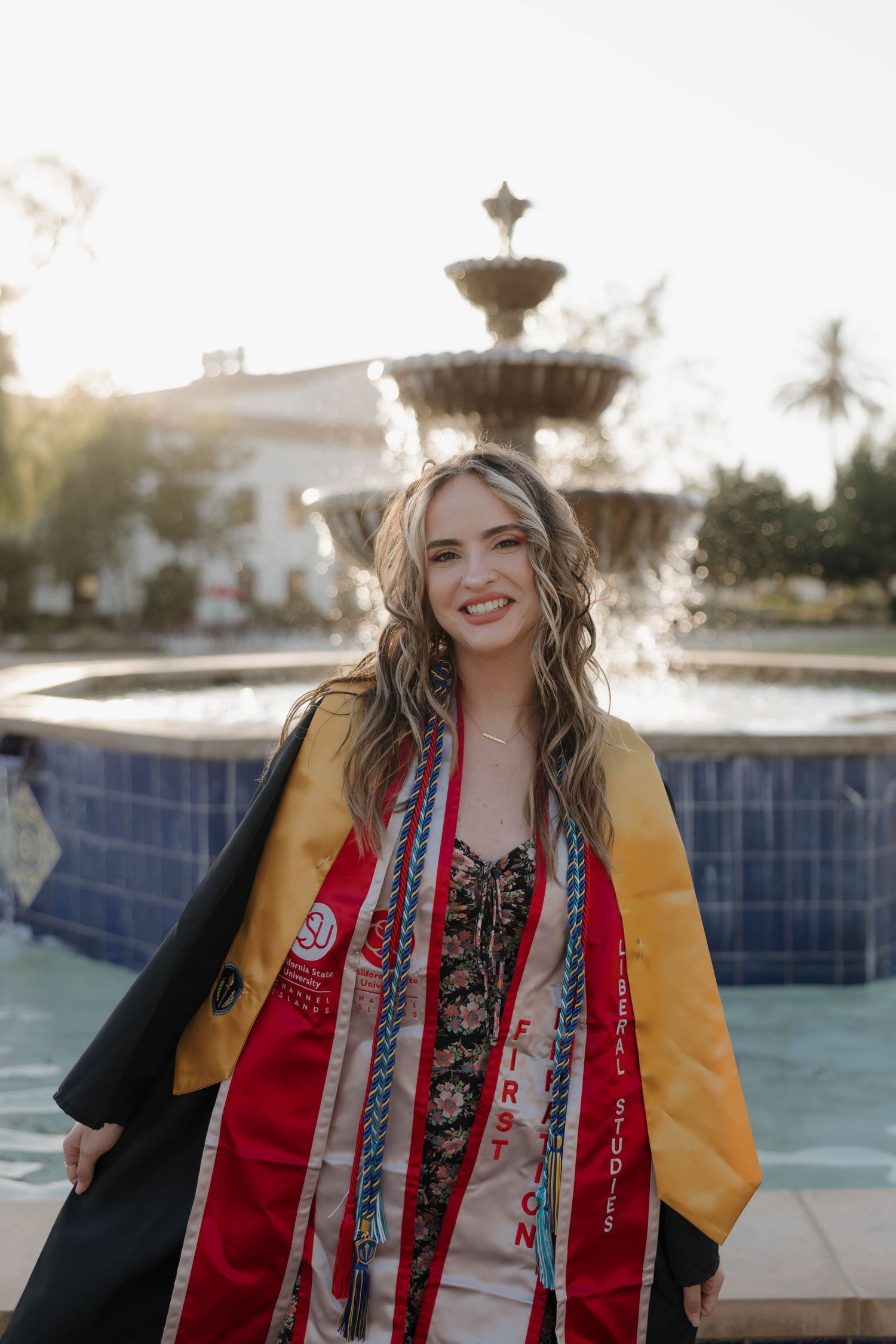 A graduation photo taken outdoors featuring academic regalia and stoles in front of a decorative fountain at sunset.