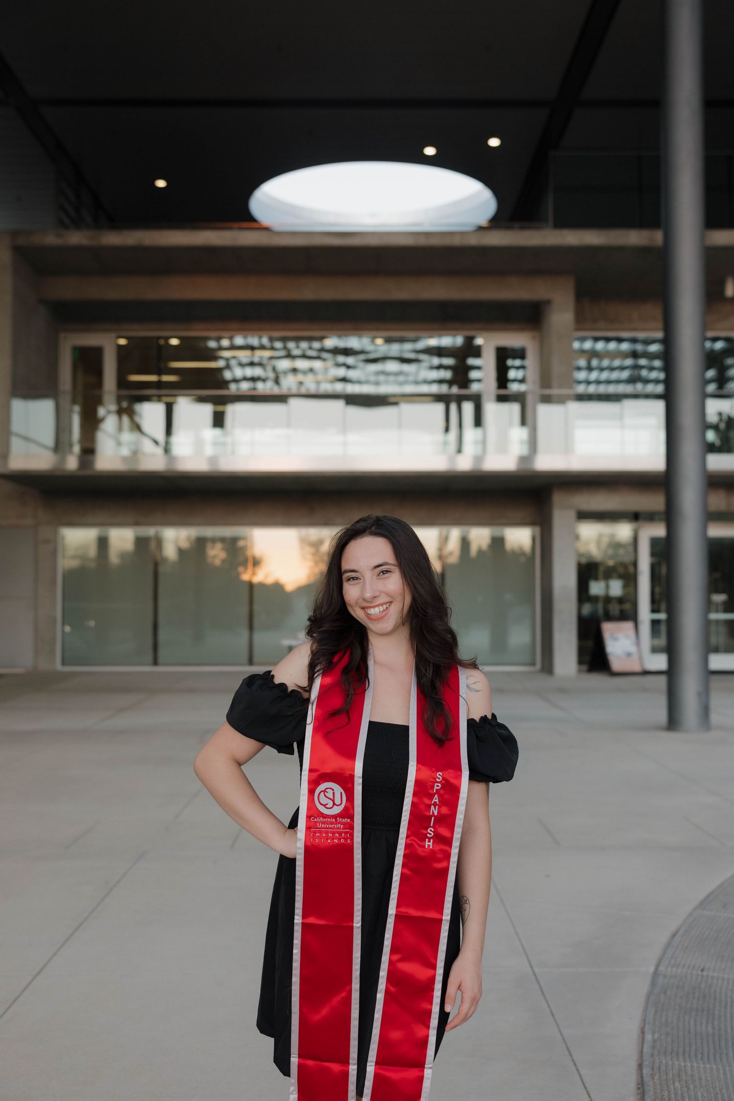 College graduate wearing black dress and red graduation stole stands in front of modern building at sunset.