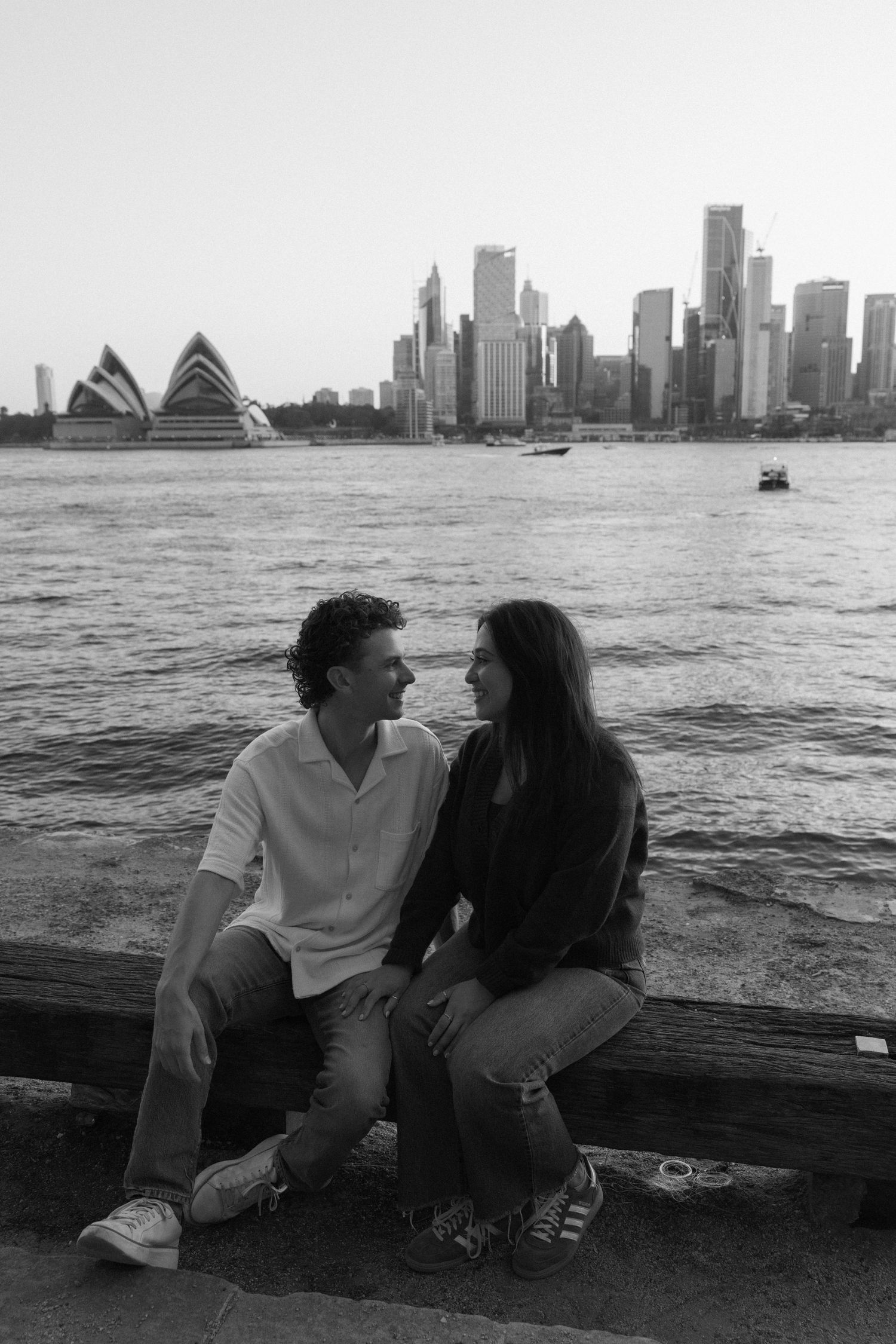Couple sitting together by Sydney Harbour with Opera House and city skyline in black and white.