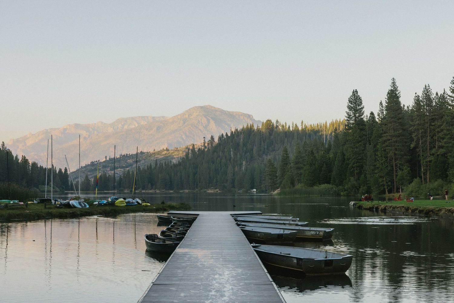 Serene mountain lake at sunset with a wooden dock, rowboats, tall pine trees, and a majestic peak in the background.