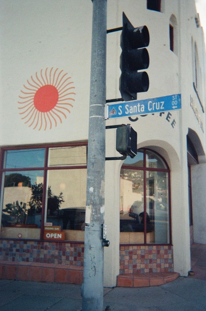Corner of S Santa Cruz street with a traffic light pole, open coffee shop, and sun mural on white building facade.