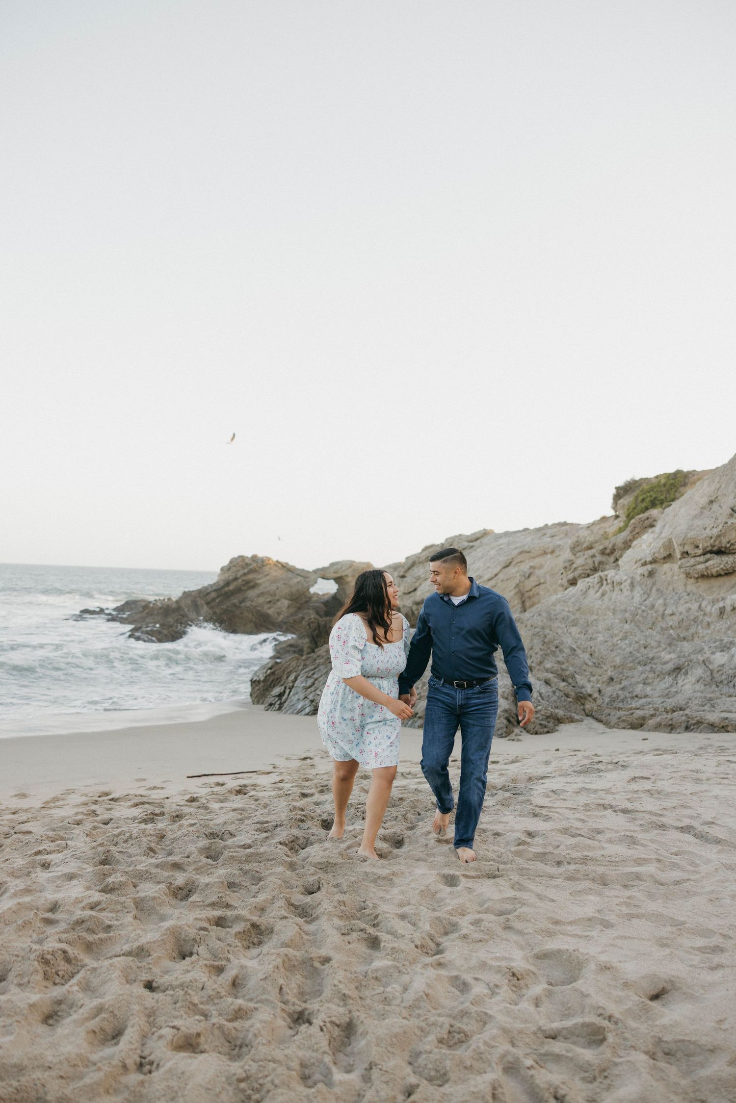 Couple holding hands and walking on a sandy beach with rocky cliffs and ocean waves in the background.