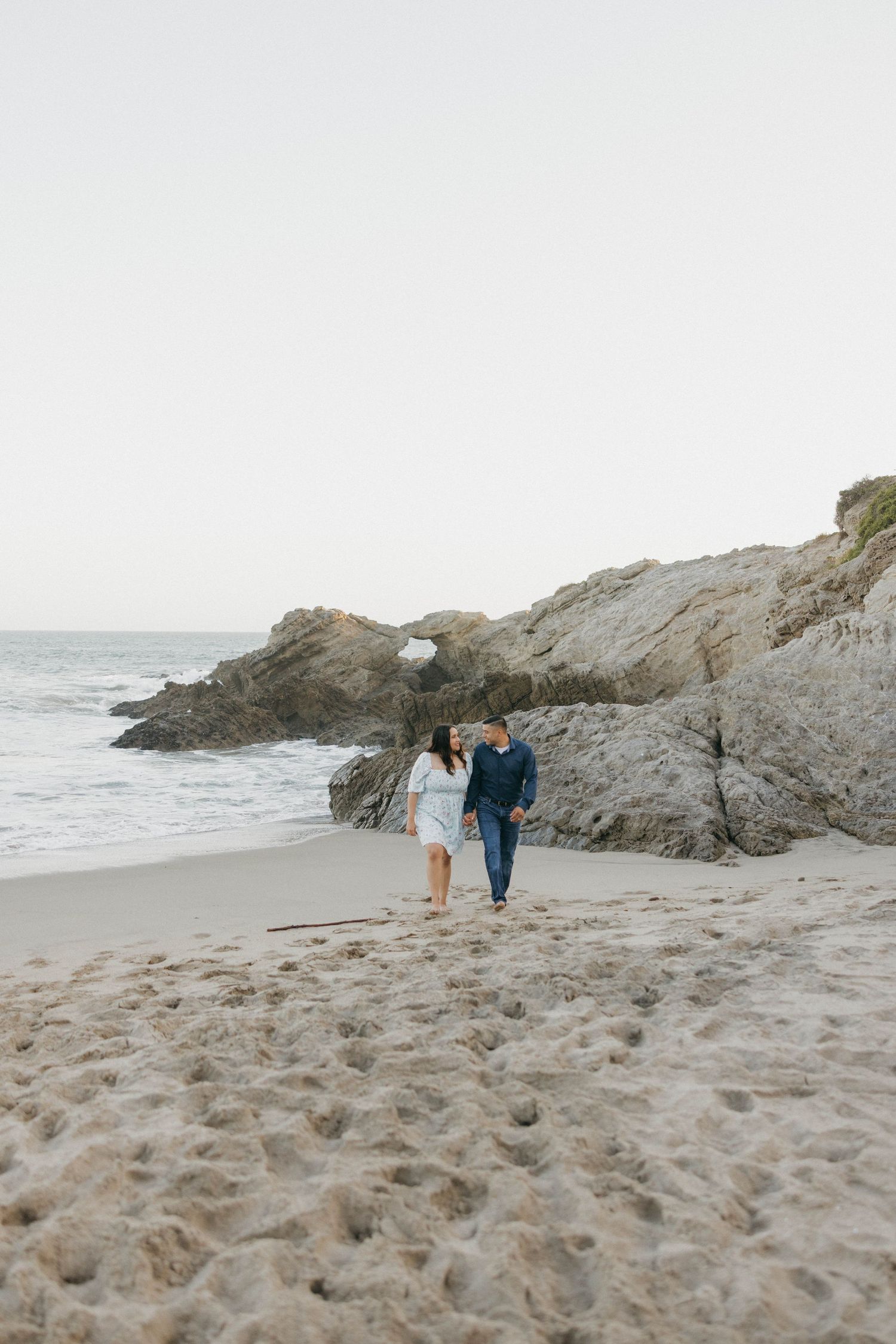 A couple walking hand in hand along a scenic beach with rocky cliffs and crashing waves at sunset.
