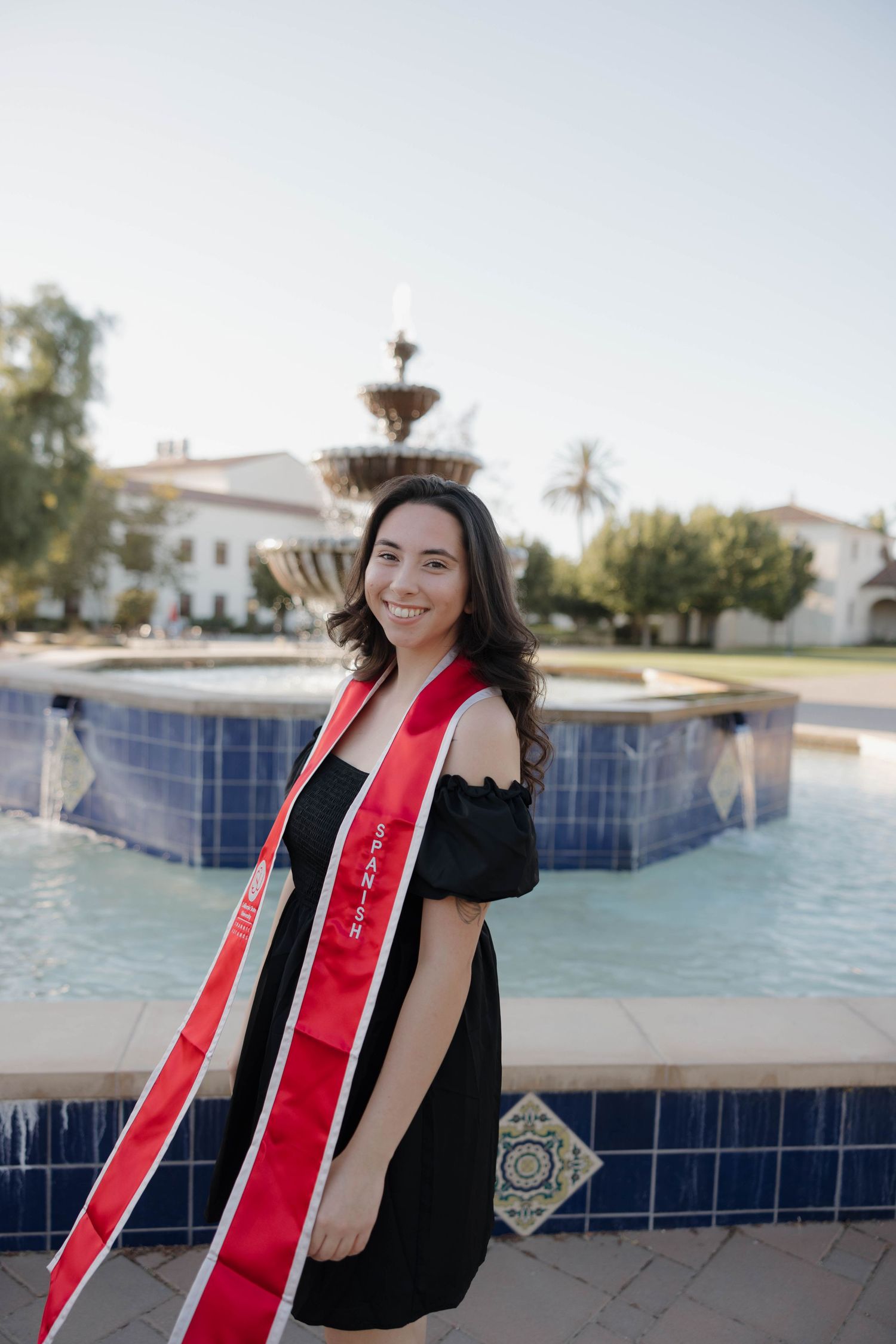 Graduate wearing a black off-shoulder dress and red graduation stole poses near a blue-tiled fountain on a sunny day.