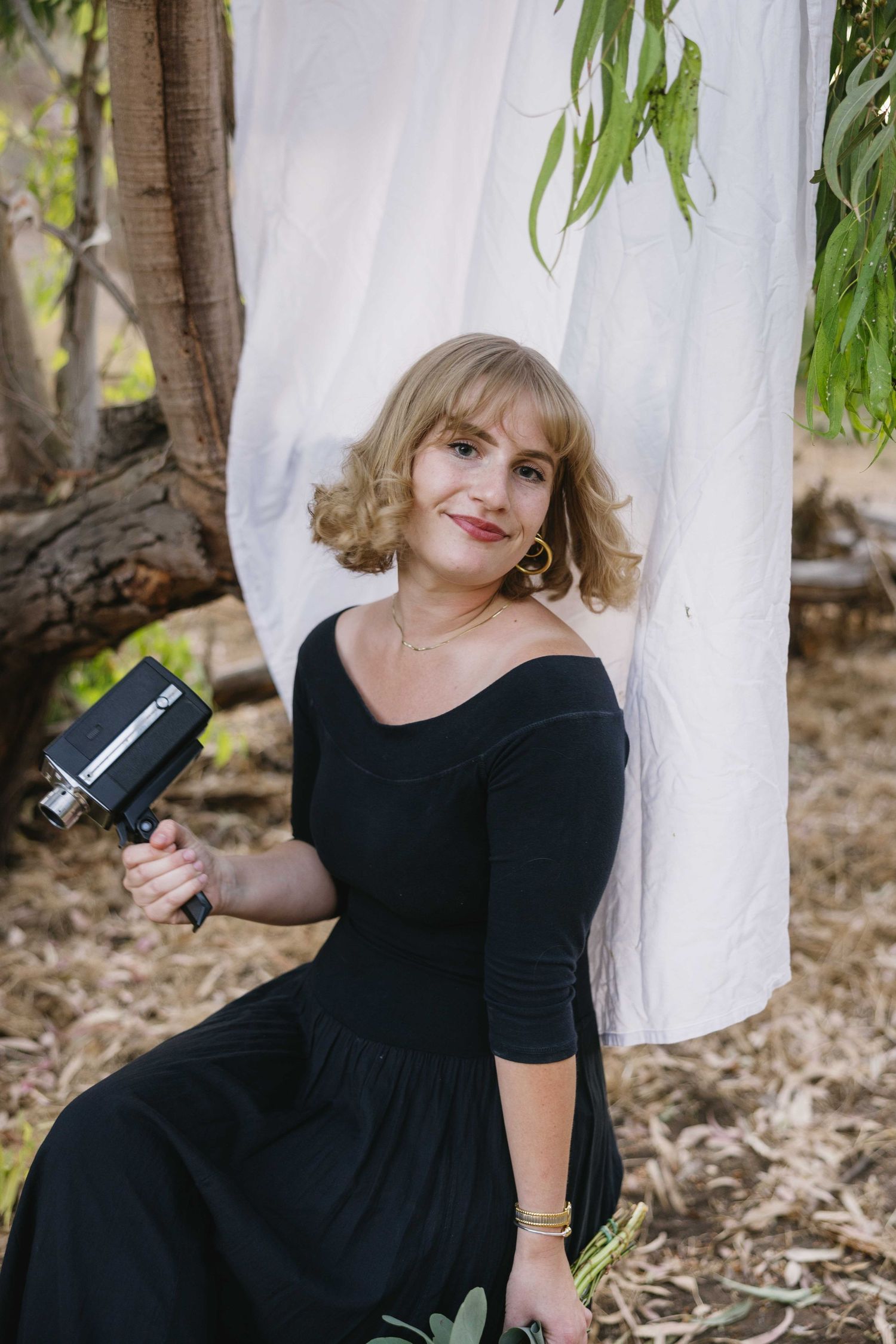 Woman in black dress poses outdoors against white backdrop with vintage camera and hanging eucalyptus leaves.