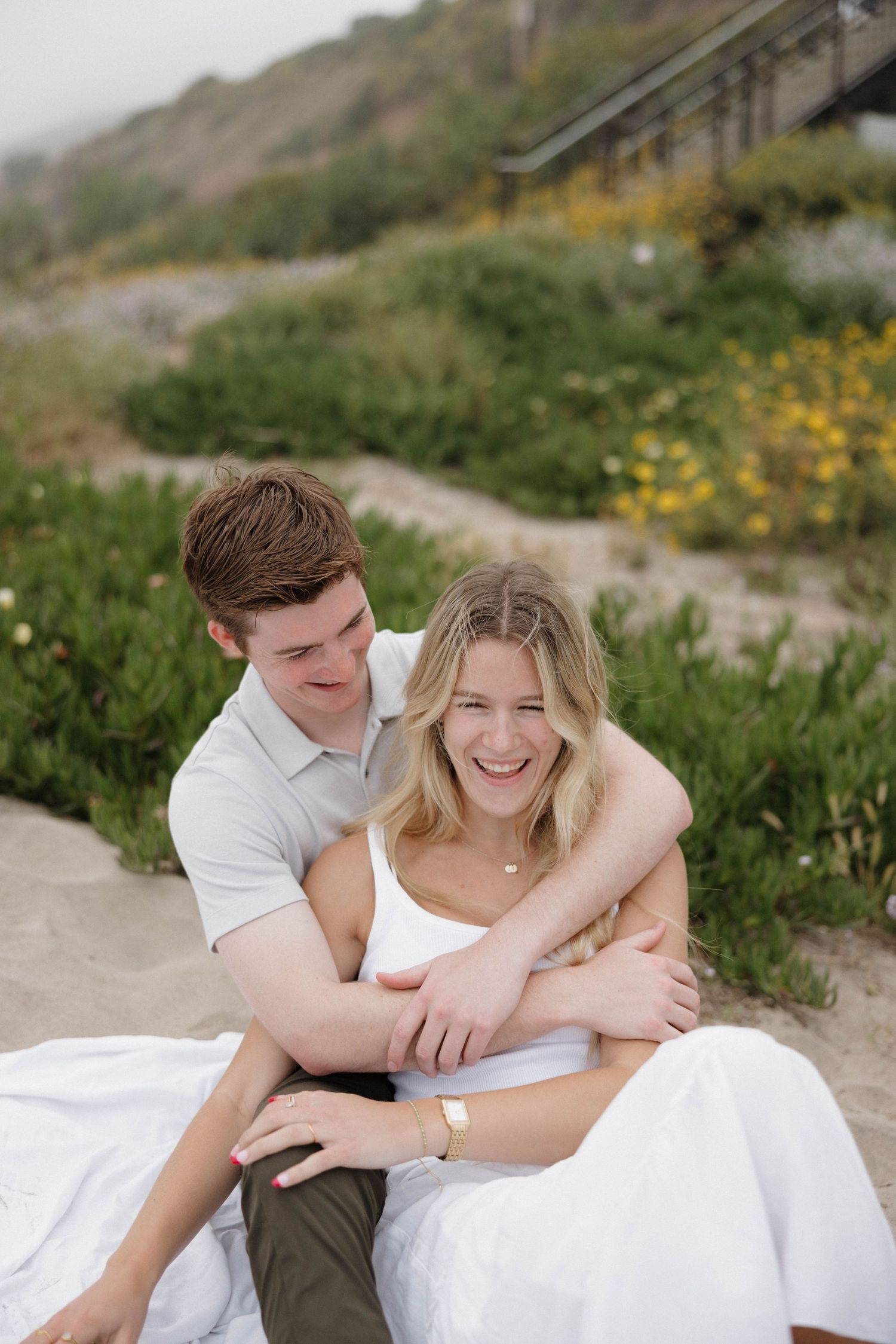 Couple embracing on sandy ground surrounded by coastal vegetation.