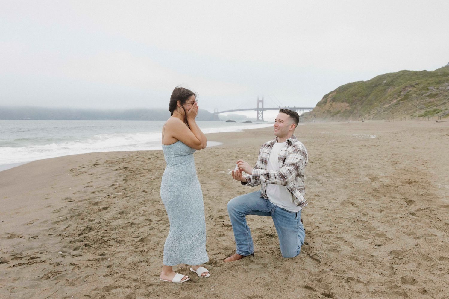 A romantic proposal moment on a sandy beach with the Golden Gate Bridge visible in the background on a misty day.