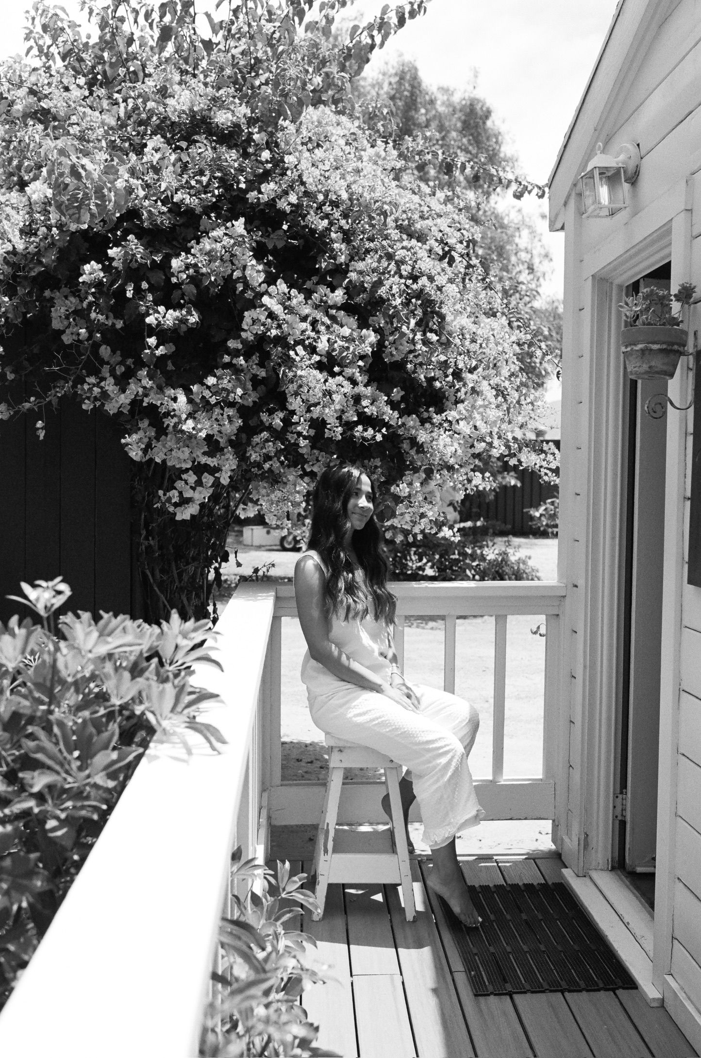 Woman in white dress sitting on stool on sunny porch with blooming trees in black and white photo.