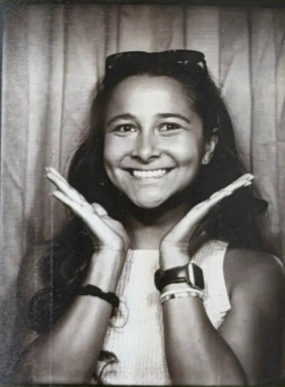Black and white photo booth portrait of a smiling young woman with sunglasses on head, hands framing her face.