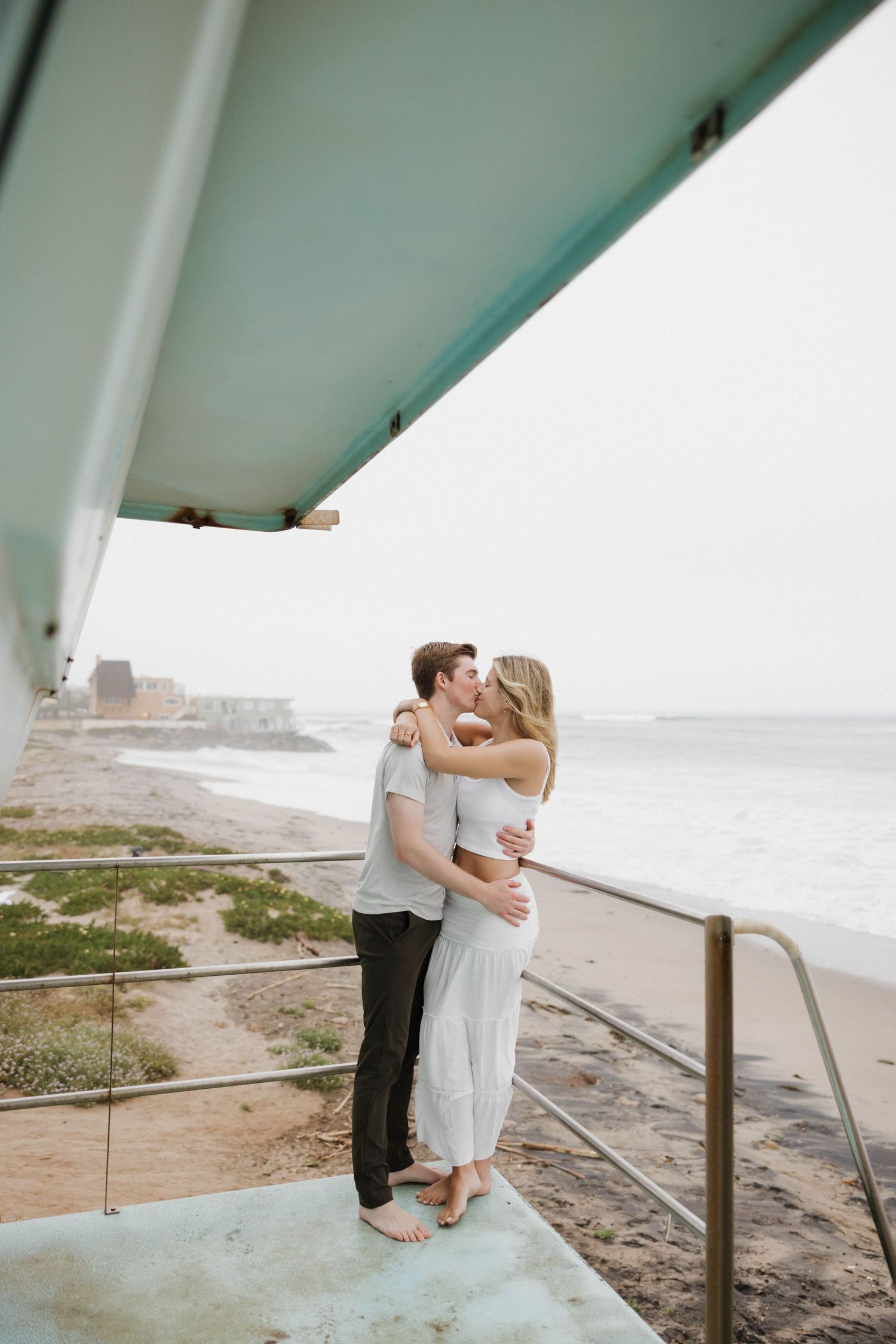 A romantic couple embraces on a seaside overlook during an overcast day at the beach.