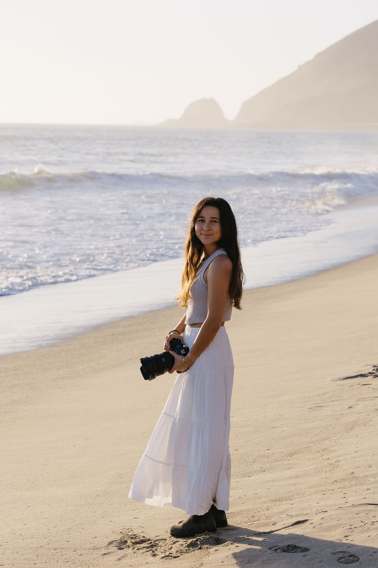 A photographer in a white dress captures photos on a sandy beach during golden hour.