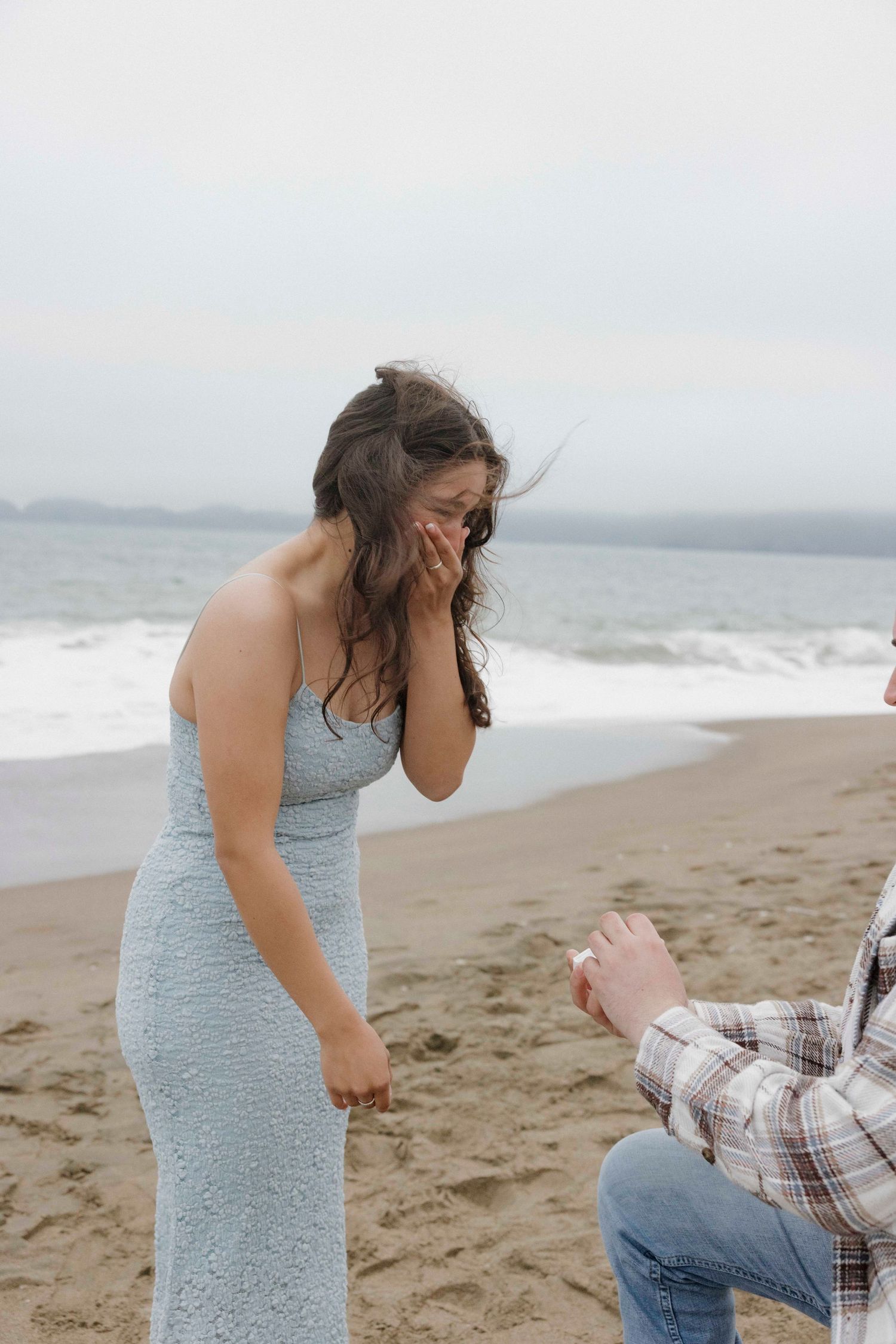 A memorable moment captured on a misty beach as someone proposes while kneeling in front of a woman in a pale blue dress.