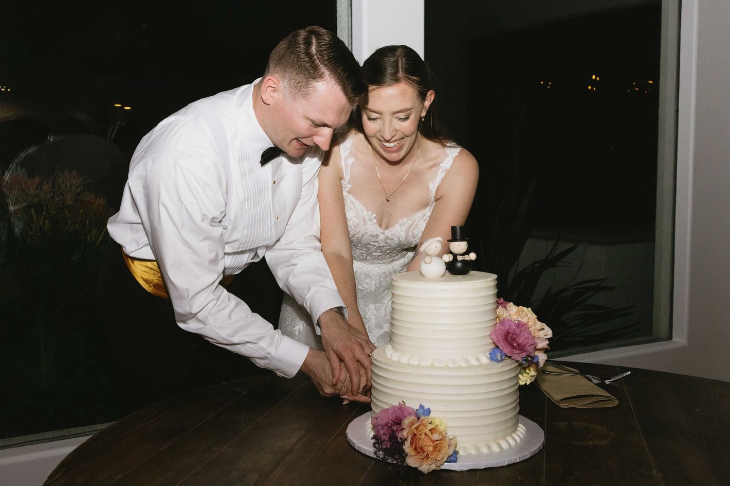 A couple cuts into their elegant two-tiered white wedding cake decorated with fresh flowers during their reception.