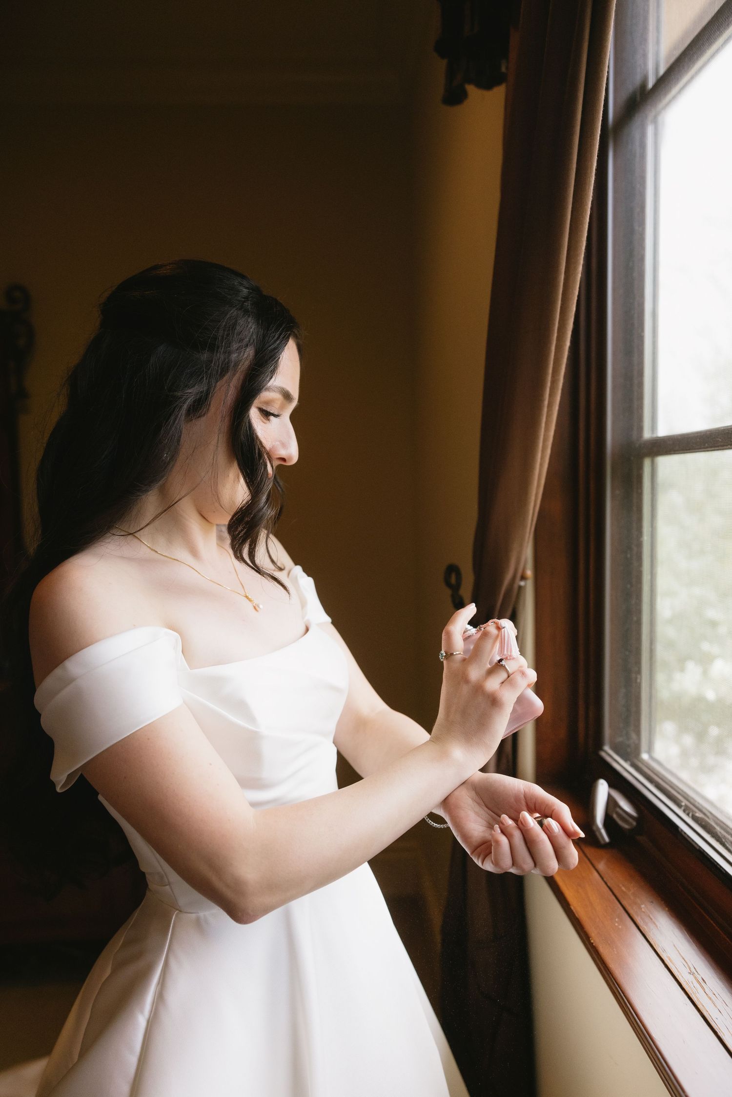 Bride in white off-shoulder gown applying perfume by a window in soft natural light.