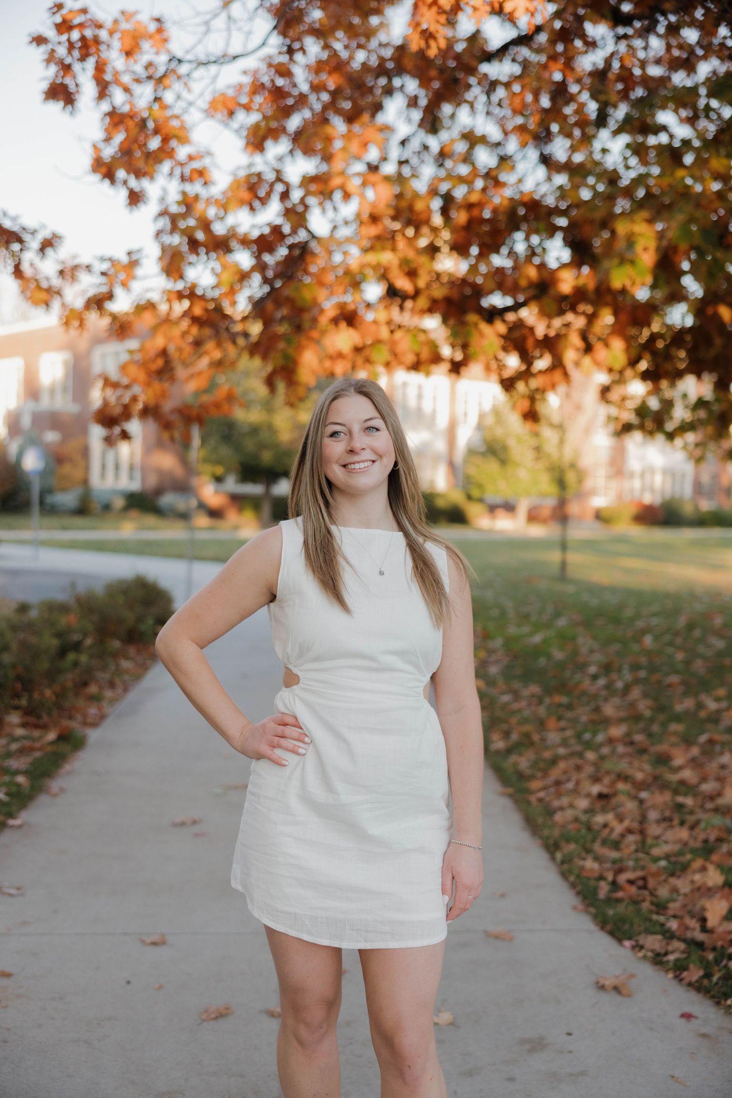 Young woman in white sleeveless dress poses on autumn sidewalk with colorful fall foliage and buildings in background.