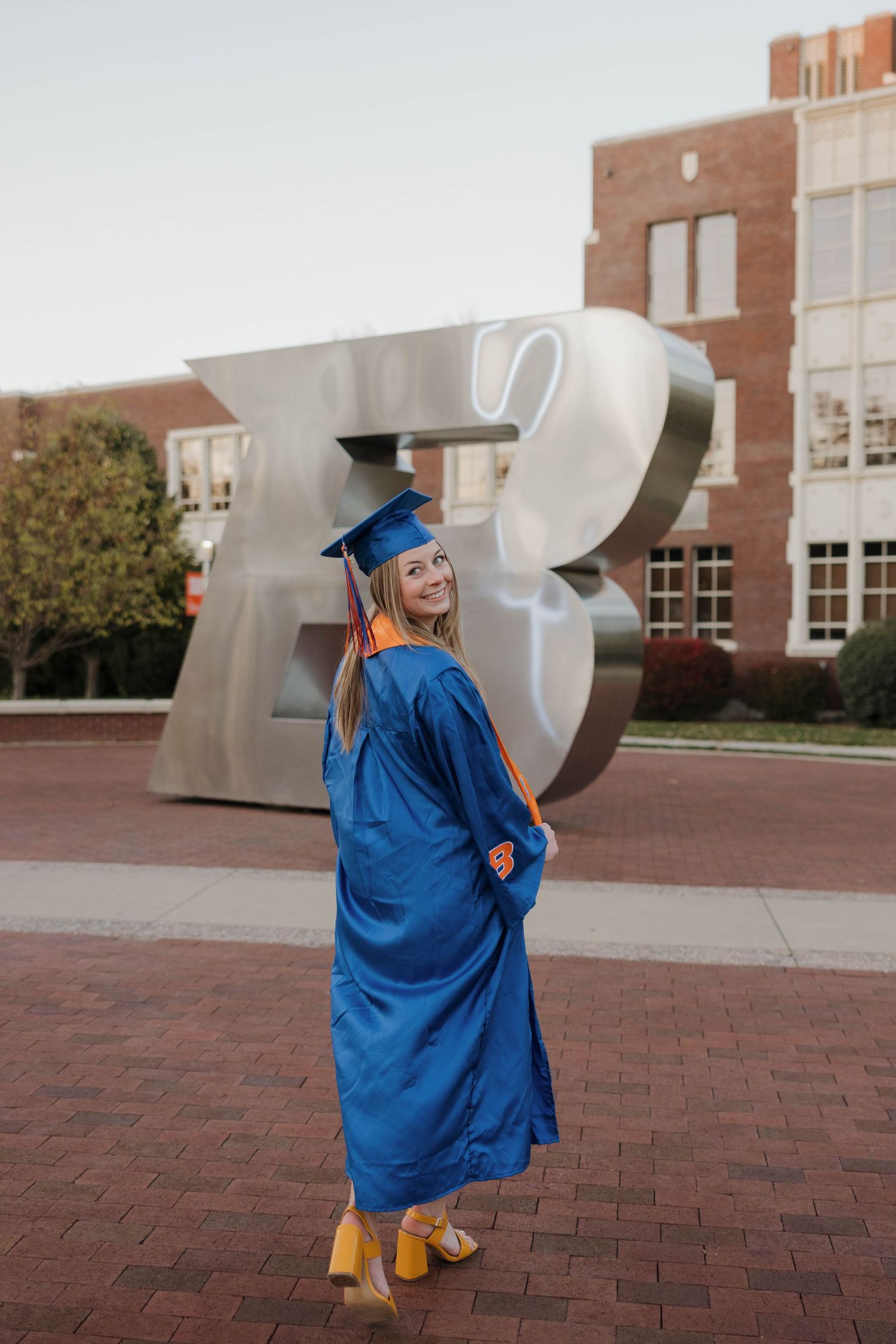 Graduate in blue cap and gown poses by large metal B sculpture on brick campus courtyard.