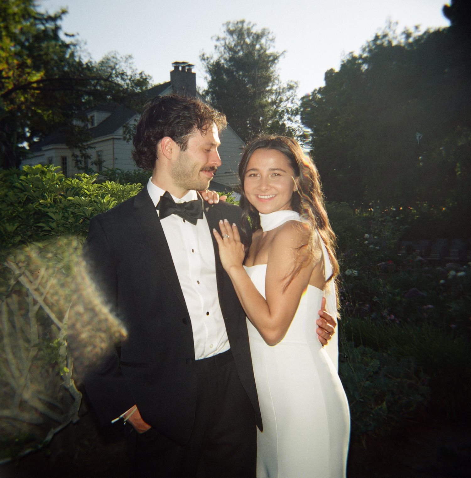 Couple in wedding attire smiling outdoors, groom in tuxedo and bride in white gown, surrounded by lush greenery.