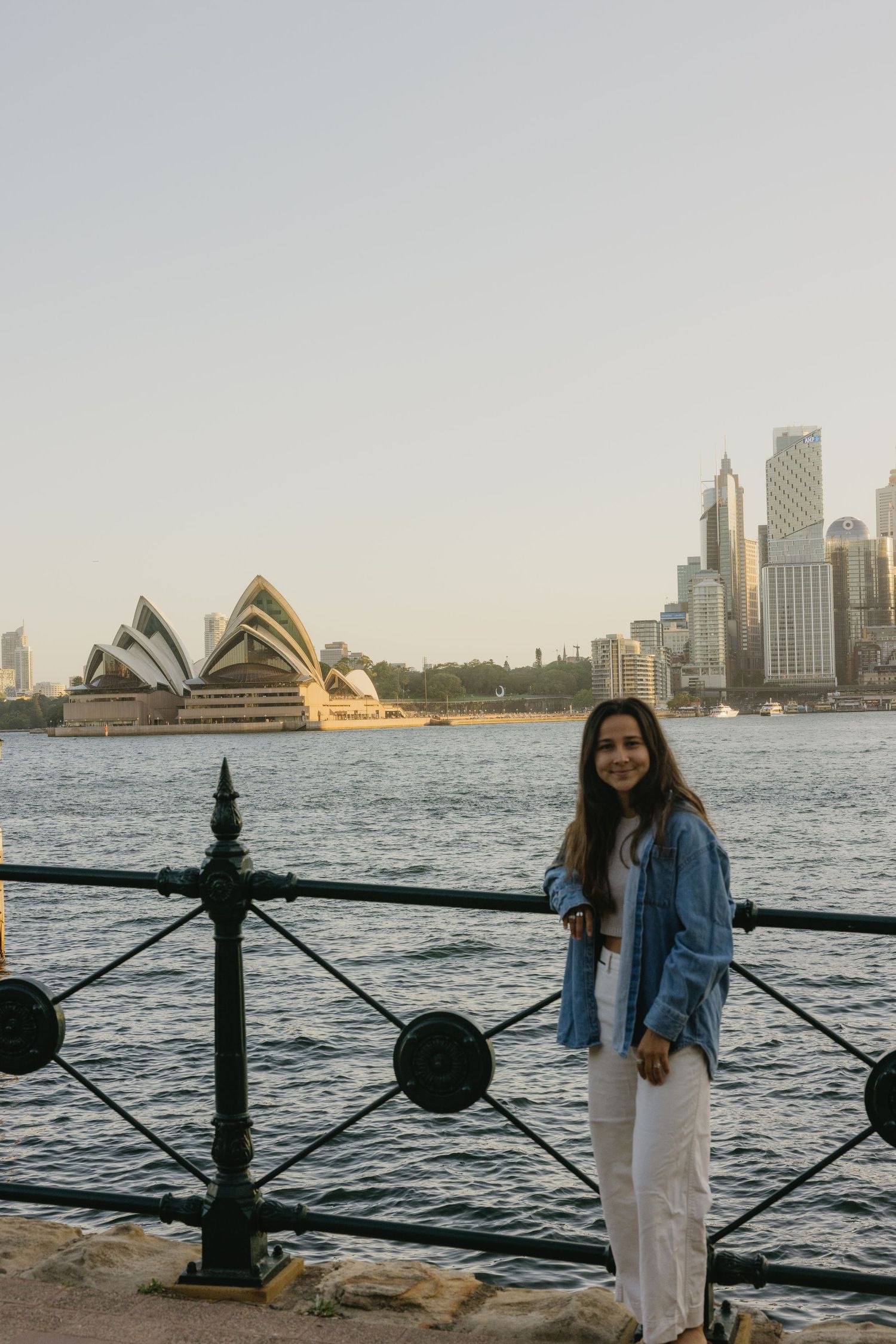 Woman posing by Sydney Harbour with iconic Opera House and city skyline at golden hour.