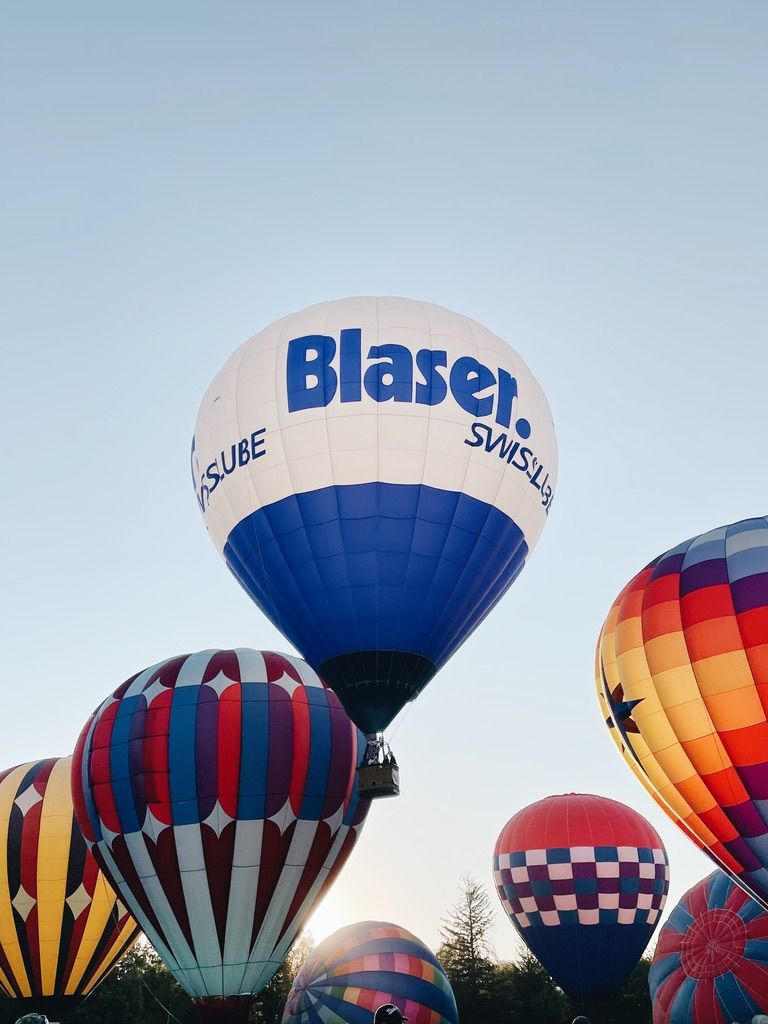 Colorful hot air balloons fill the sky at a festival, with a prominent white Blaser Swiss balloon in the foreground.
