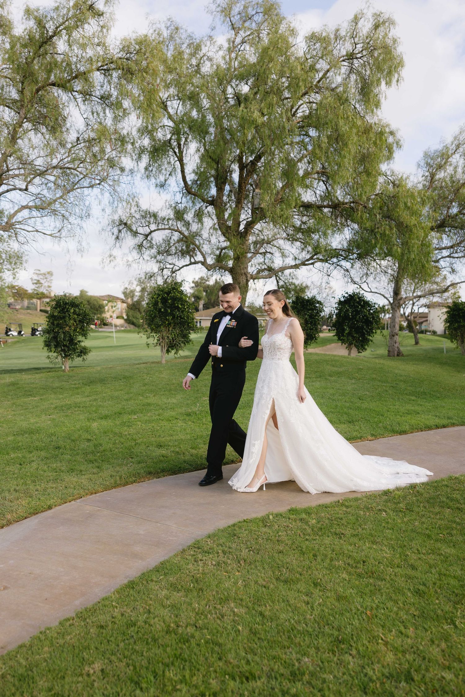 A couple walks along a winding path through a lush green lawn with a large willow tree in the background.