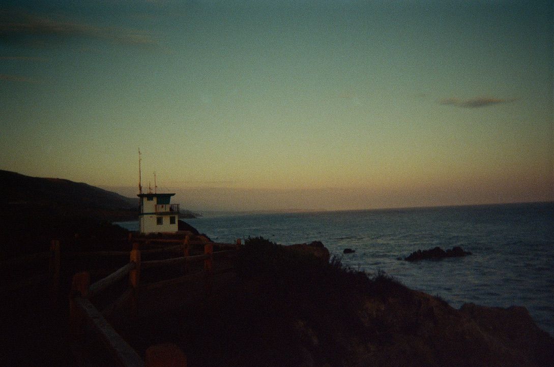 Lighthouse silhouette against dusky ocean sunset with rocky coastline.