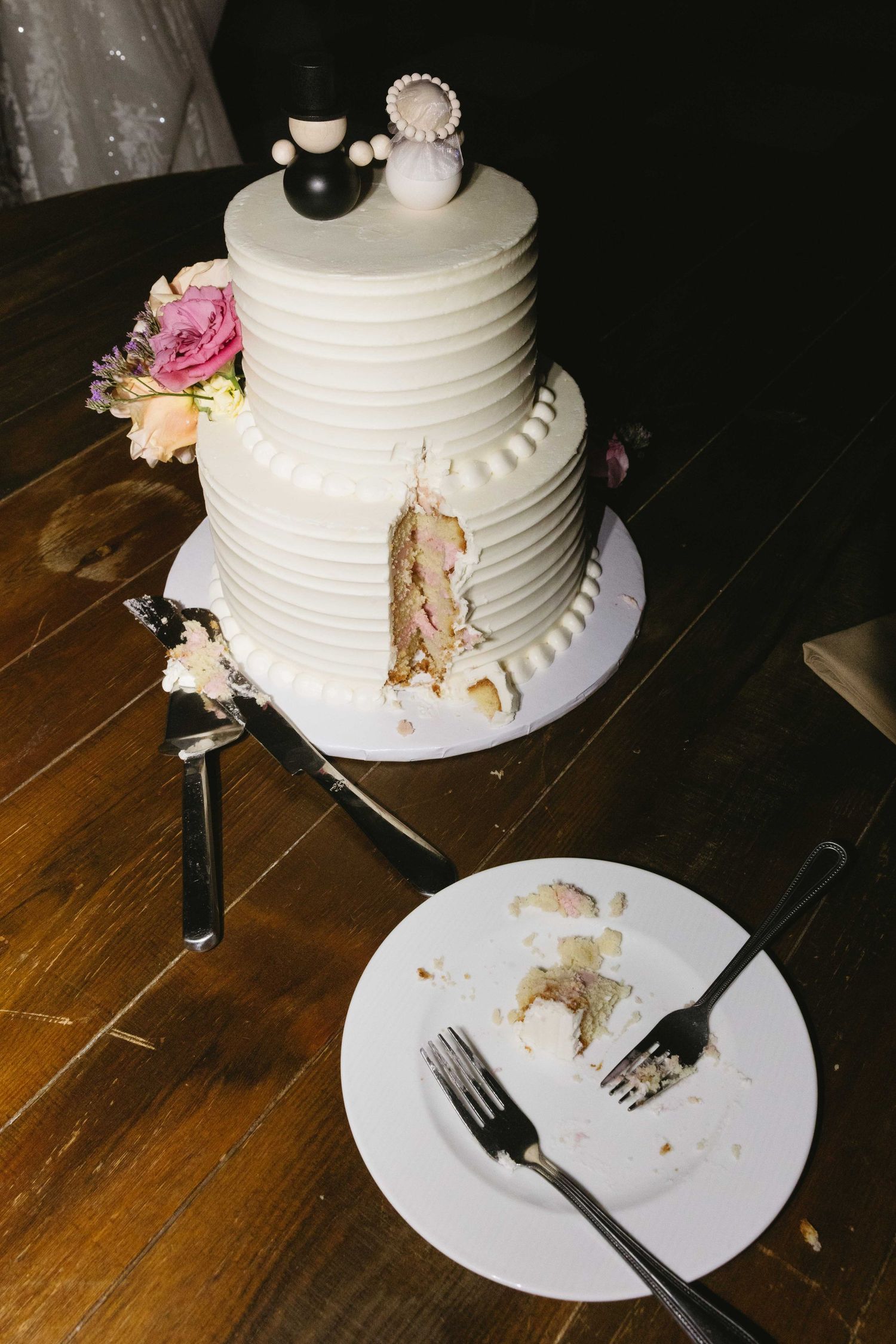 White tiered wedding cake with cake toppers sits on wooden table next to plate with crumbs and used utensils.
