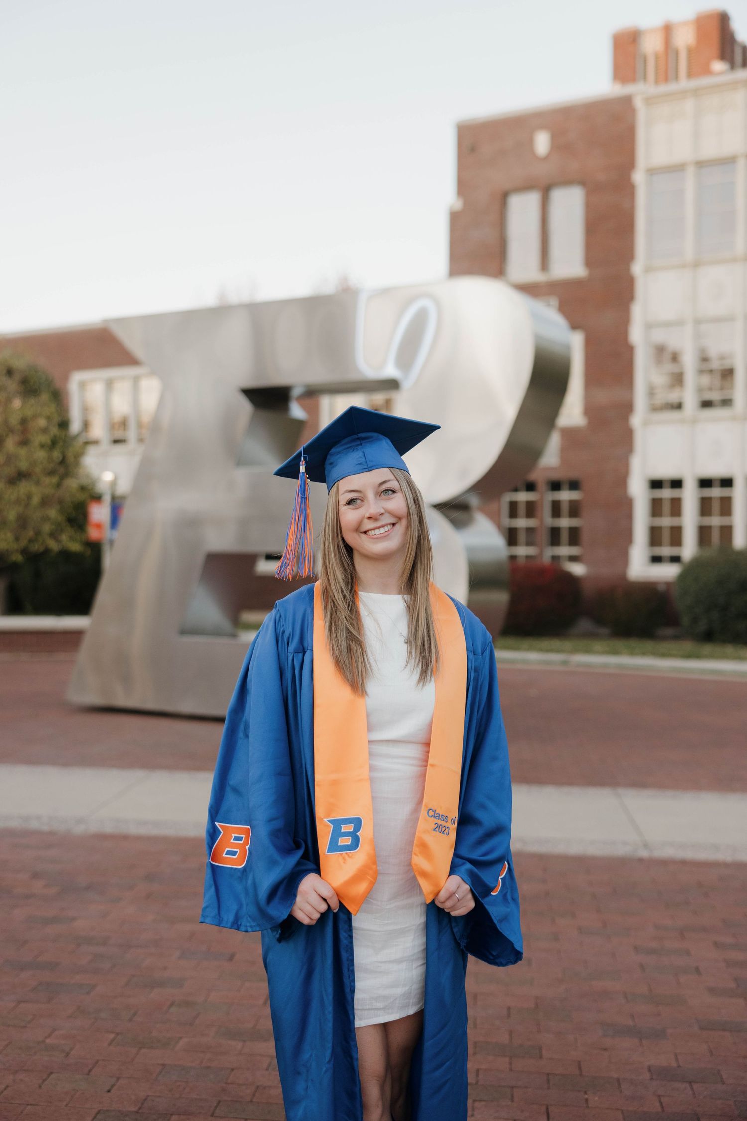 Graduate in blue cap and gown with orange stole stands smiling in front of modern campus sculpture on brick plaza.