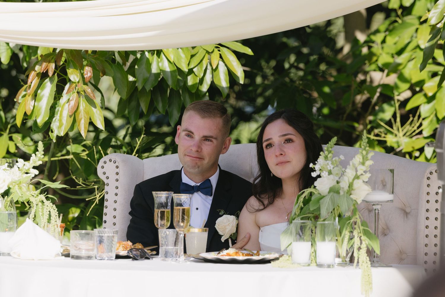 A couple sits together at an elegant outdoor wedding reception table decorated with greenery and white linens.