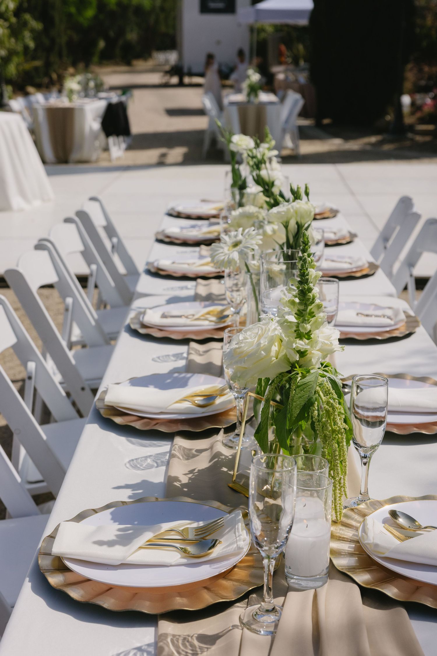 Elegant outdoor table setting with white floral centerpieces and gold charger plates for a summer wedding reception.