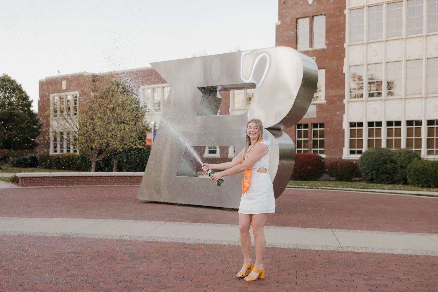 Person in a white dress poses playfully with a large metallic sculpture outside a brick building.