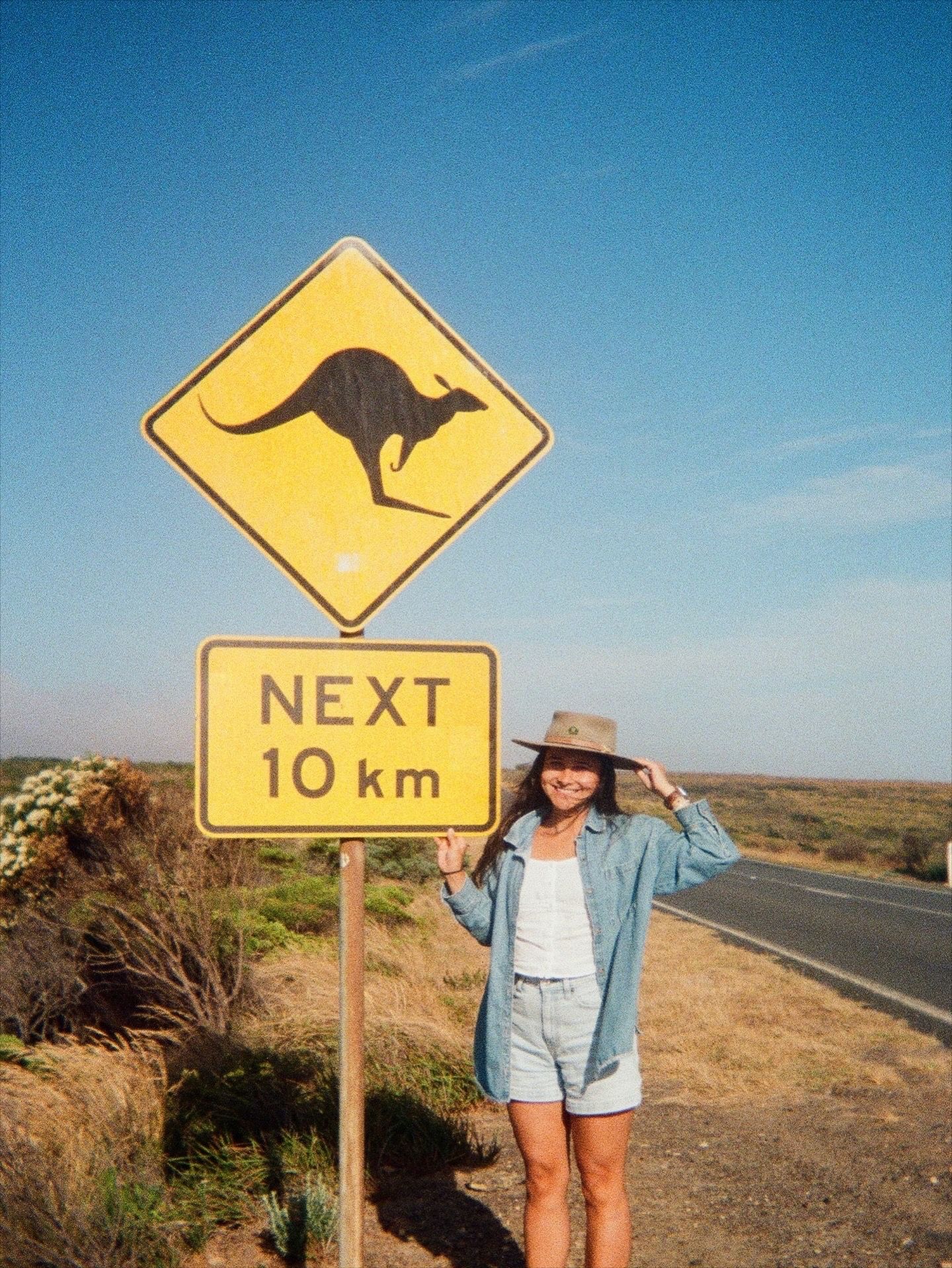 Woman posing next to Australian kangaroo warning road sign reading Next 10 km on a sunny outback highway.
