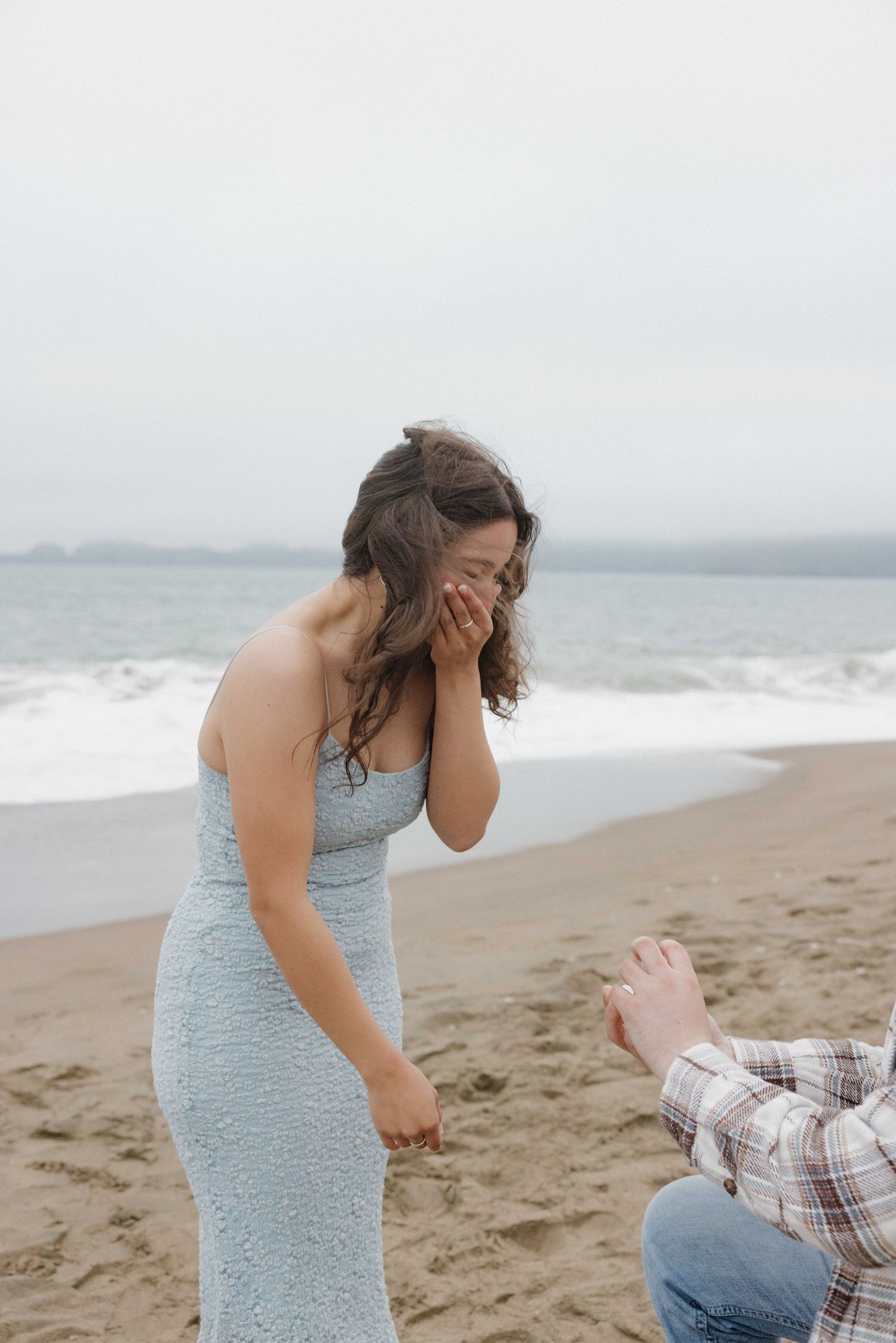A person in a light blue dress reacts with surprise as someone proposes on a misty beach.