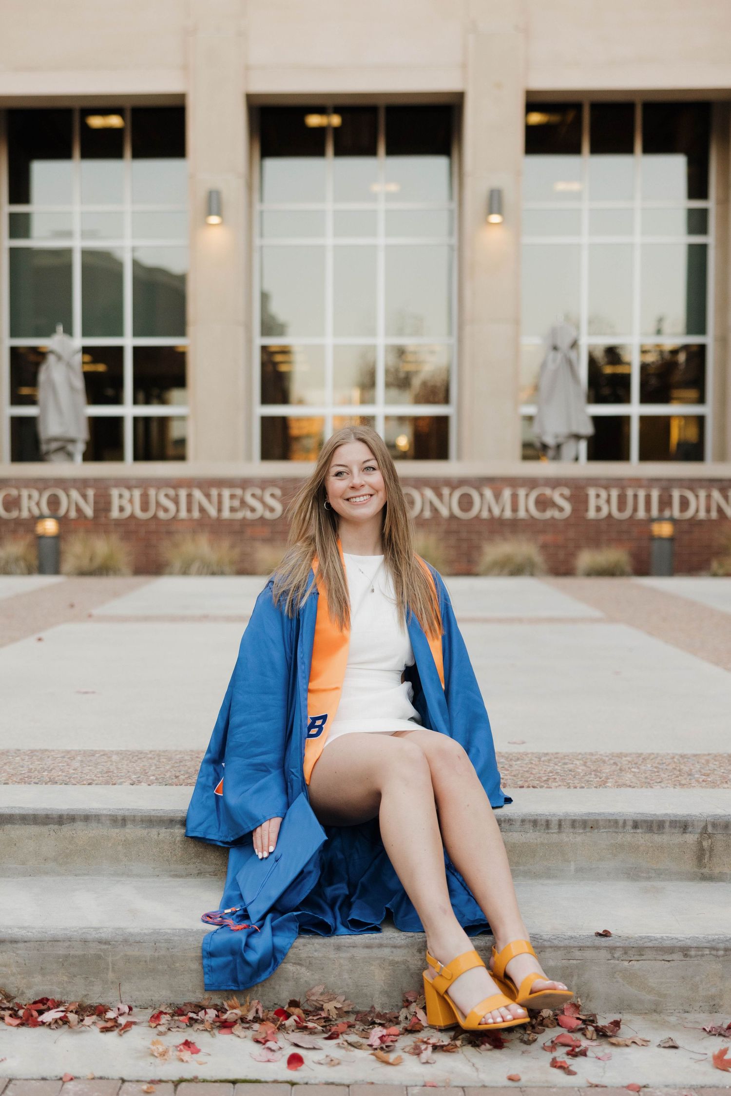 A graduate in a blue gown sits on steps outside an economics building in yellow shoes and a white dress.