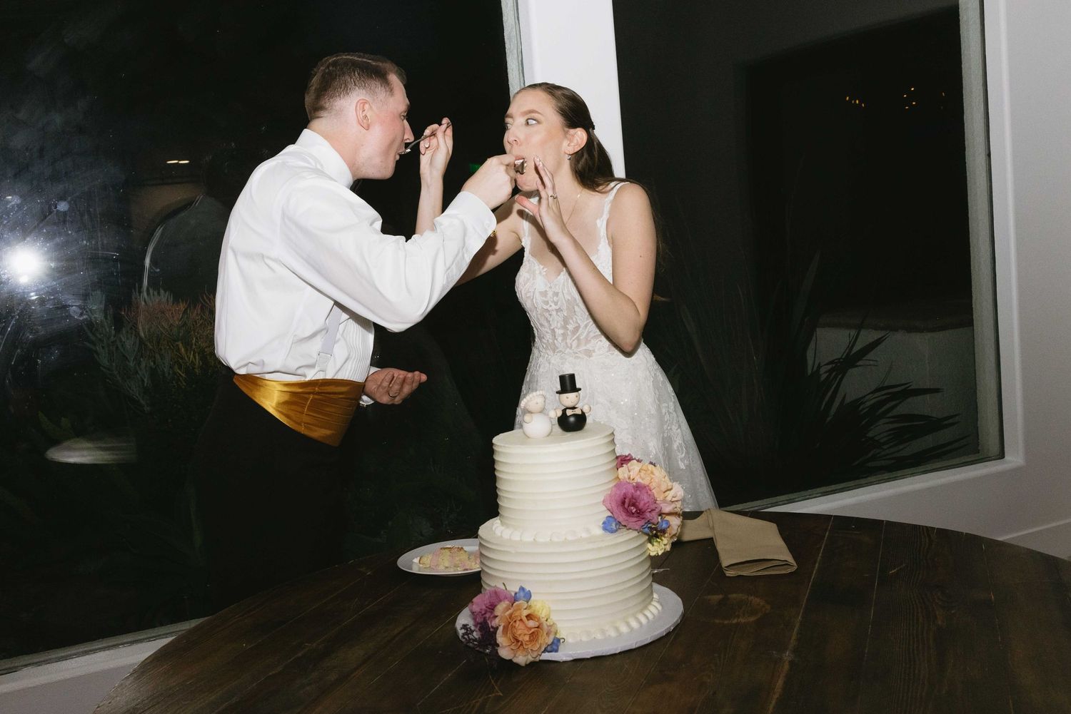 A couple shares wedding cake at their reception as they feed each other a bite in front of their floral decorated tiered cake.