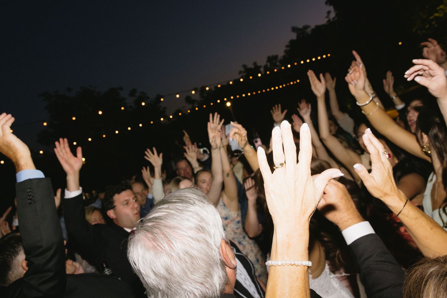 Crowd of wedding guests raising hands on outdoor dance floor with string lights at night.