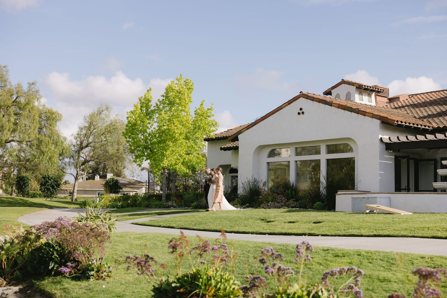 A panoramic view of a luxury white Spanish-style home with lush green landscaping and rolling hills in the background.